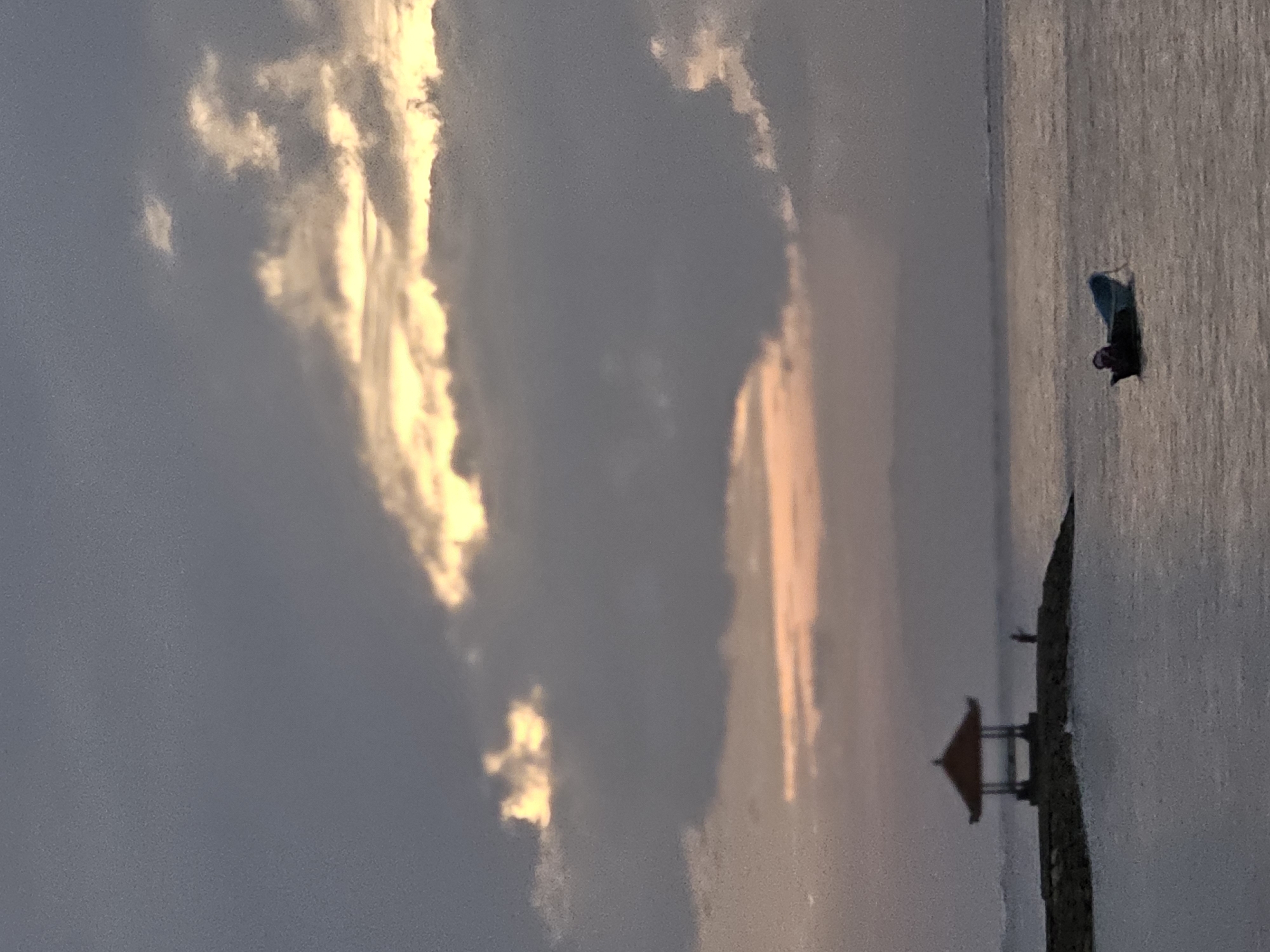 The image shows a serene seascape at what appears to be sunrise or sunset. The sky is filled with dramatic clouds, with patches of golden light breaking through, suggesting the sun is low on the horizon. In the foreground, the calm water reflects the muted colors of the sky. To the left, a stone jetty extends into the water, topped by a small, traditional Balinese pavilion or gazebo with a dark roof. A lone figure stands near the pavilion, seemingly observing the view. Further out on the water, a small blue boat is moored, with what appears to be a person seated inside. The overall atmosphere is peaceful and contemplative. This scene is likely captured at Sanur Beach in Denpasar, Indonesia, a popular location known for its sunrise views and distinctive beach structures.