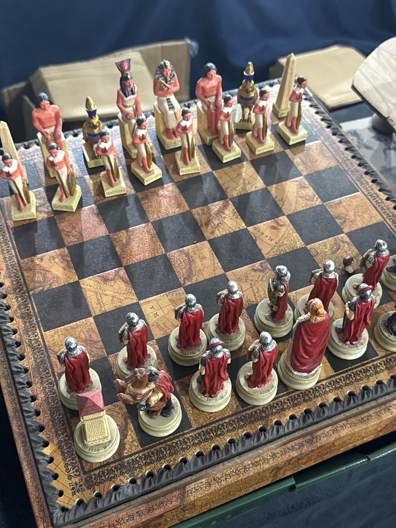 The image shows a close-up view of an Egyptian-themed chessboard set up on a patterned board. The pieces appear to be made of a resin or plastic material and are painted in various colors, including red, white, gold, and brown. The white pieces depict figures in ancient Egyptian attire, such as pharaohs and queens, while the black pieces represent Roman soldiers. Some pieces are standing, while others are in various poses. The chessboard itself has a map-like design with a grid of black and brown squares. The board is placed on a dark surface, possibly a table. The lighting suggests an indoor setting. There is no indication of the city of Tbilisi, Georgia, or any specific location within it.
