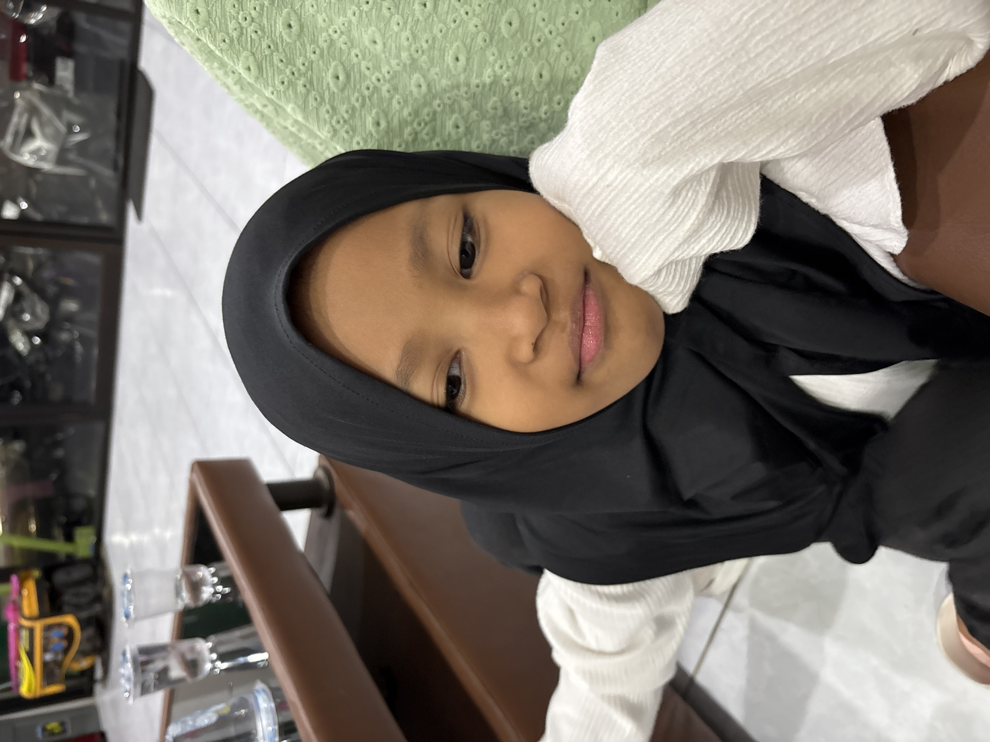 The image, captured in Kaliwates, Indonesia, features a young girl indoors. She has a light-brown complexion and is wearing a black hijab that covers her head and neck, paired with a white top made of textured, crinkled fabric. The girl is looking directly at the camera with a gentle, slight smile, conveying a calm and content expression. Her left arm is raised, with her hand resting softly against her face or ear, while she appears to be leaning on a brown wooden surface, possibly a table or a chair's armrest, which occupies the lower and right foreground.
In the mid-ground, to the left of the girl, several clear glasses or cups containing a transparent liquid are visible on the brown surface. The background is partially blurred, showing an indoor setting. Behind the girl, a light green fabric with a patterned, possibly embroidered, texture is visible. To the far left, a dark, reflective area with indistinct shapes suggests shelving or a display case holding various items, hinting at a commercial establishment or a well-furnished public space. No visible text is present in the image.