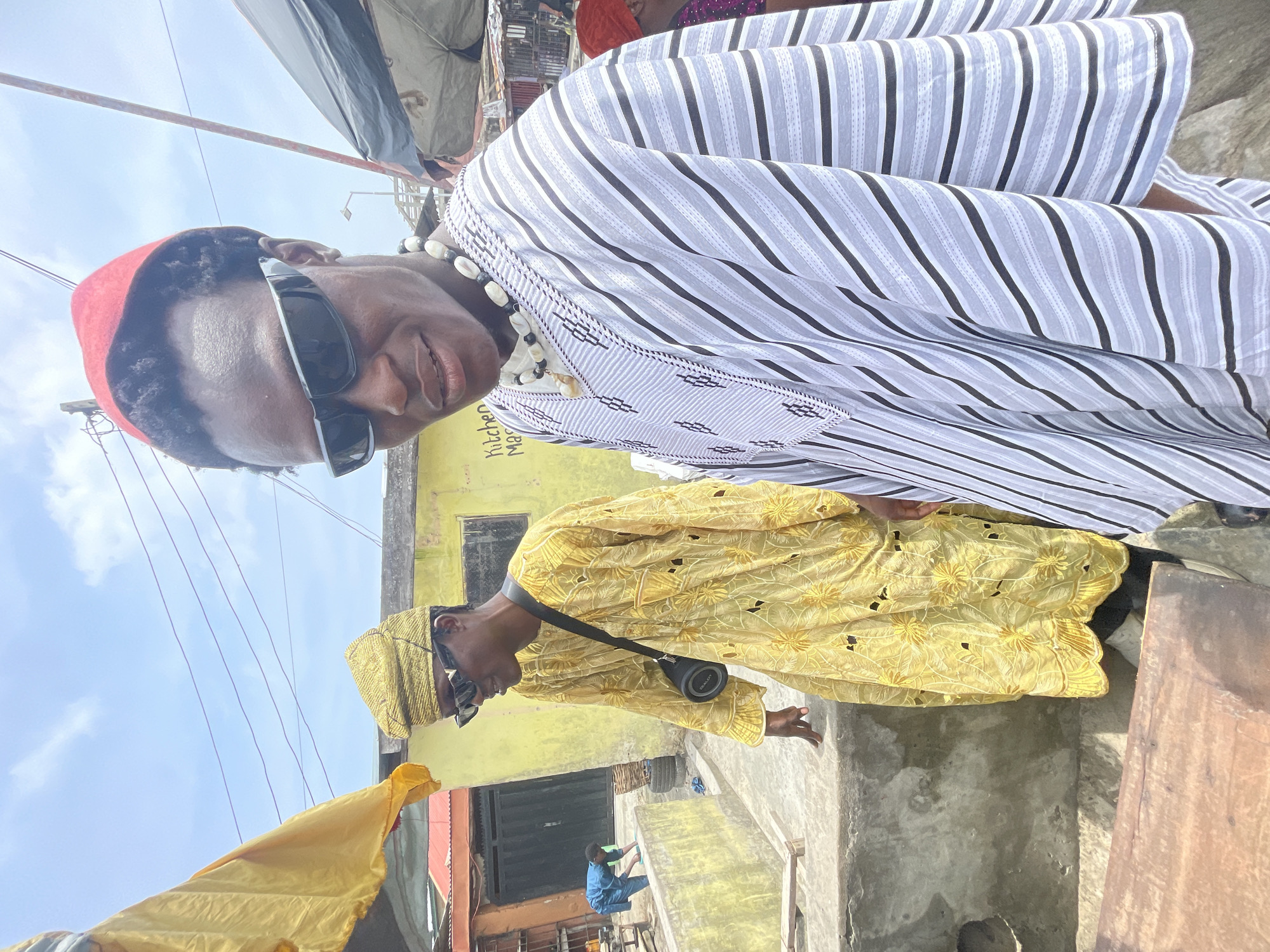 Two men stand outdoors in Ado, Nigeria. The man in the foreground wears a striped robe, sunglasses, and a red cap. A necklace of white shells adorns his neck. Behind him and slightly to the right, another man wears a yellow robe and head covering, sunglasses, and carries a black cylinder on a strap. A yellow building is visible behind them. Below, a small person dressed in blue works near the ground. The sky is partially visible. The environment appears to be a street with various items and structures present.