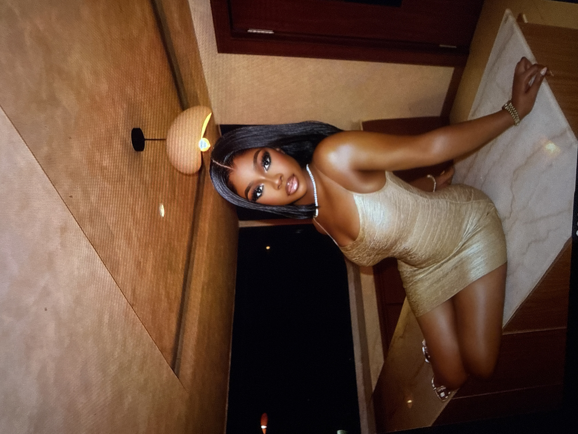 A woman with dark skin and long, dark hair is sitting on a marble-topped surface, possibly a counter or table, in a dimly lit room. She is wearing a shimmering, short, gold dress with spaghetti straps and a diamond necklace. Her makeup is elaborate, with winged eyeliner and glossy lips. She is wearing a bracelet on her right wrist and rings on her fingers. The background is dark, suggesting it is nighttime. A modern pendant light hangs from a sloped, wood-paneled ceiling. The scene appears to be an indoor setting, possibly a restaurant or lounge, with wooden elements and a warm, intimate ambiance. There is no visible indication of the city of Abuja, Nigeria in the image.