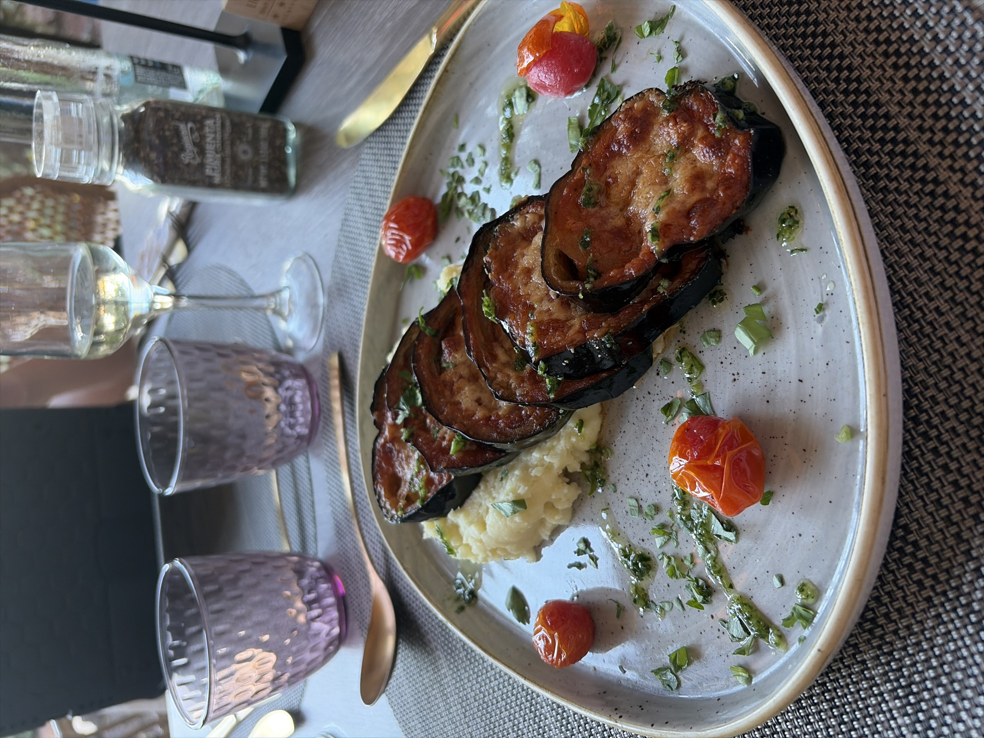 A close-up shot captures a meticulously plated meal on a table, likely in a restaurant or dining setting. The central subject is an oval-shaped plate holding several slices of roasted or grilled eggplant, appearing golden-brown with dark skin, arranged over a bed of creamy mashed potatoes. The dish is garnished with small, bright red and orange cherry tomatoes, chopped green herbs, and drizzled with a light green sauce or olive oil.
To the left of the plate, a clear wine glass stands beside two textured, opaque glasses in a purple-pink hue. Further left and slightly behind, a clear glass seasoning grinder is visible, containing dark specks, likely black pepper. Text on the grinder partially reads "PIMIENTA NEGRA" (Spanish for black pepper) and "NET. WT. 3.5 OZ". A golden-colored knife rests on the placemat near the plate, and a matching spoon is partially visible further down. The plate itself is light grey with a visible speckled texture and a beige rim, sitting on a dark grey, woven placemat. The overall presentation suggests a well-prepared meal in Antigua, Spain.
