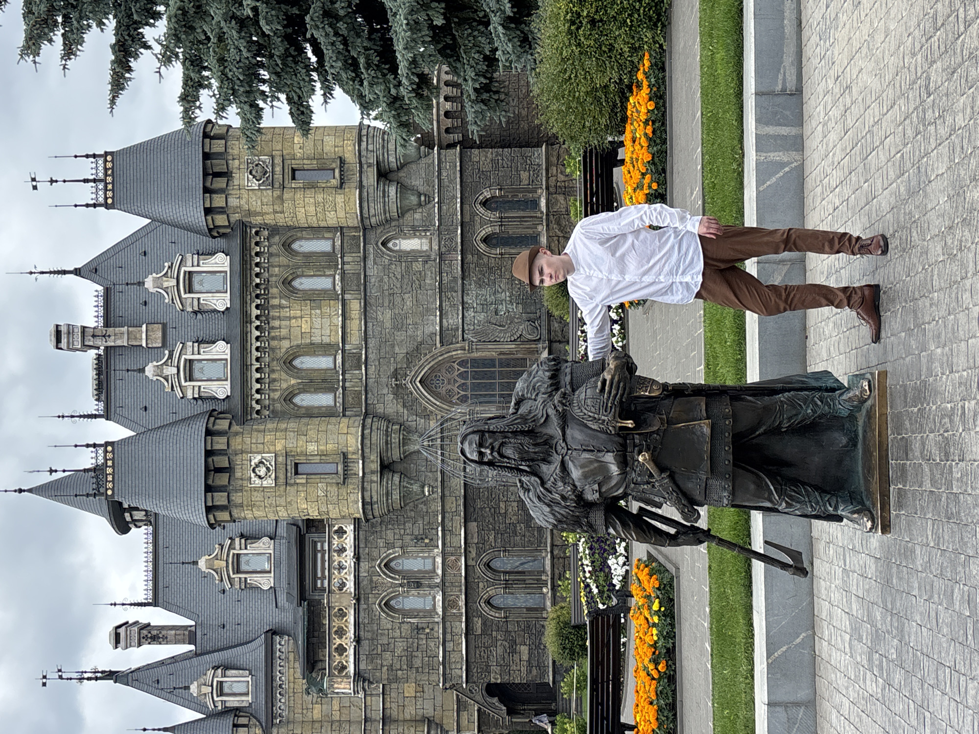 A young man in a white button-down shirt, brown pants, and a brown fedora stands next to a large bronze statue of a man with a beard. The statue is dressed in historical armor and holds a helmet in its right hand and an axe in its left. The man is looking to his left, with his right arm extended and touching the statue's shoulder. The background is dominated by a large, gothic-style stone building with multiple towers and arched windows. In front of the building, there are flowerbeds filled with orange and white flowers, benches, and a large evergreen tree on the right. The scene is set in the city of Khryashchevka, Russia, where this building is known as the "Fairy Tale Castle" and is part of the "X-Park" amusement area.