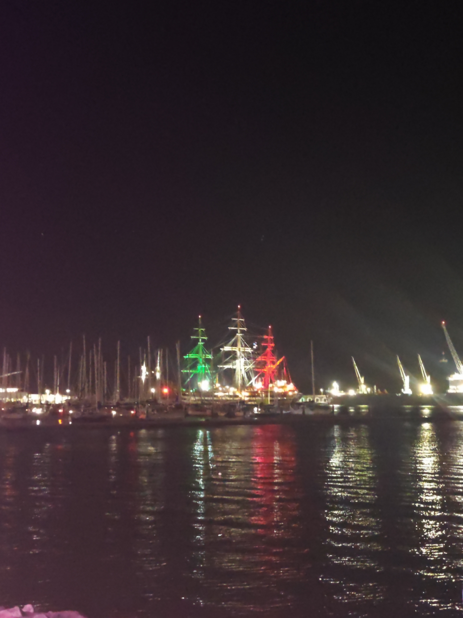 This is a nighttime photograph of the Port of Ponta Delgada, Portugal. In the foreground, the dark water of the harbor reflects the lights from the ships and the shore. The middle ground is dominated by a fleet of docked sailboats and tall ships. One large sailing vessel in the center is illuminated with green lights, and another to its right is lit with red lights, suggesting a festive or national theme. In the background, several large cranes stand illuminated against the dark sky, indicative of port operations. The overall scene depicts a busy, well-lit harbor at night.