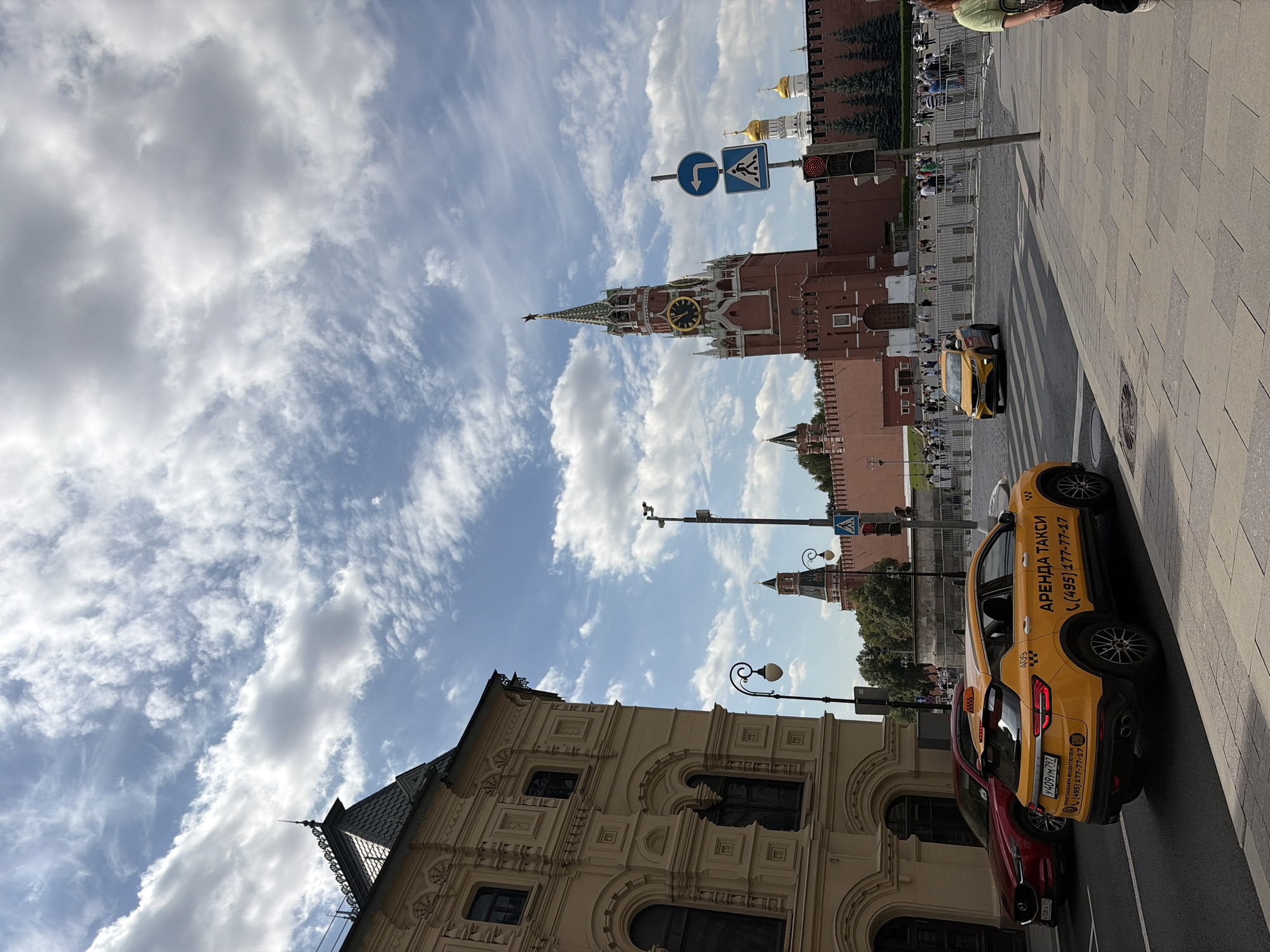 This is a medium-angle, eye-level shot of a street scene in Moscow, Russia, with the iconic Kremlin and St. Basil's Cathedral in the background. The sky is partly cloudy, with a mix of bright sunshine and cumulus clouds.
In the foreground, on the left, is the corner of a large, ornate, tan-colored building with arched windows and decorative carvings. To the right of this building, a street runs diagonally into the frame. Several vehicles are visible on the street. A red car is parked on the left side of the street. In front of it, a yellow taxi with Russian license plates and text in Cyrillic is driving away from the camera. Another yellow taxi is driving towards the camera in the middle of the street.
In the background, the red brick walls of the Kremlin are prominent, with several towers and domes visible. The Spasskaya Tower, with its clock, is the most distinguished structure. St. Basil's Cathedral, with its colorful onion domes, is partially visible behind the Kremlin walls. A crowd of people can be seen walking in front of the Kremlin, behind a metal barrier. Traffic lights and street signs are also visible. A street lamp with a distinctive curved post stands to the left of the yellow taxi in the foreground. The ground is paved with cobblestones and concrete tiles.