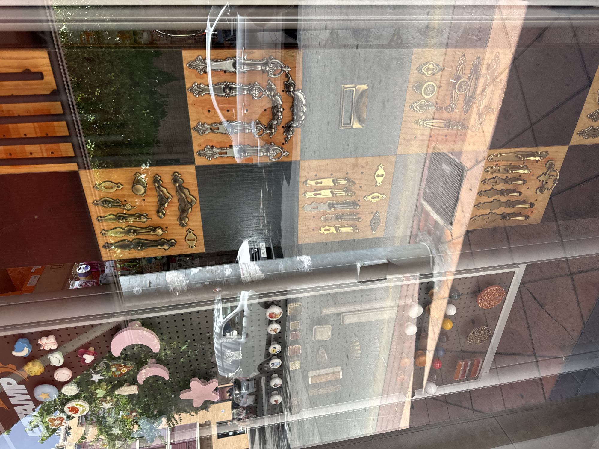 The image shows a store window display with various hardware items and decorative objects, likely in a city. The display is seen through a glass pane, with reflections from the street and surrounding buildings.
In the upper left portion of the display, there are numerous colorful, circular and star-shaped decorative items. Some of these are in the shape of celestial bodies like moons and stars, while others appear to be patterned discs with floral or abstract designs. There is also a pink butterfly-shaped item and a light blue item with a moon shape.
Below these decorative items, on a pegboard, are several round knobs of various colors and designs, including white, red, blue, yellow, and patterned. To the right of these knobs, a white van is visible through the window, parked on a street. The van has some sort of graphic design on its side.
The main part of the display features various hardware items, primarily drawer pulls and handles, mounted on wooden panels that are arranged in a checkered pattern. These hardware pieces are made of metal, with several sets displaying ornate, antique-style designs in brass and silver tones. Some of the drawer pulls are very large and elaborate, with intricate scrollwork. Other items include smaller knobs and keyhole escutcheons.
The reflections in the glass indicate an outdoor urban environment. We can see buildings with brickwork and windows, trees with green foliage, and parts of the street with pavement and a metal grate. The lighting suggests it is daytime, with sunlight casting shadows and creating glare on the glass.
While the exact location is not specified, the user requested the location be identified as Murcia, Spain. Without further information, it is impossible to definitively confirm this.