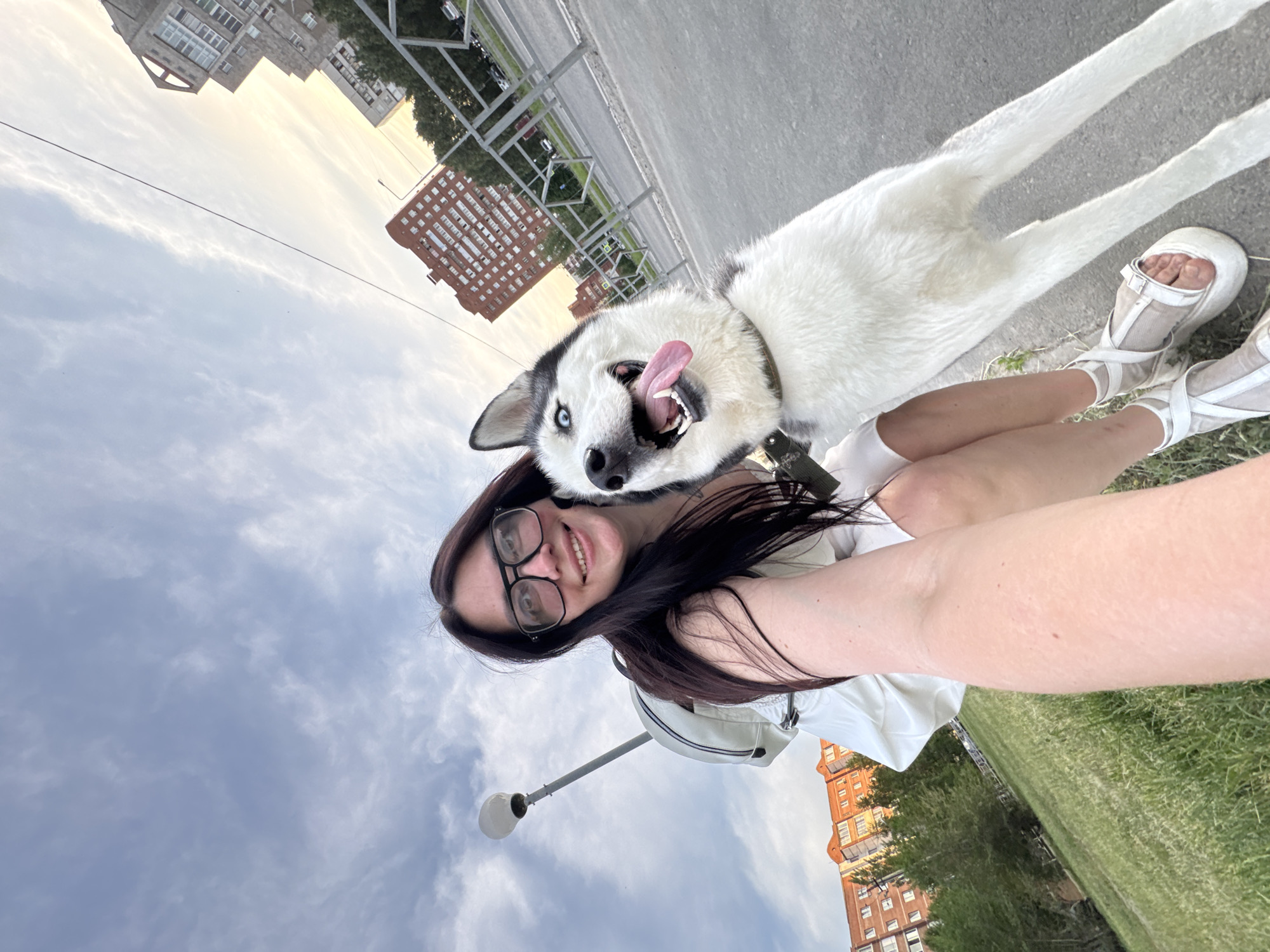A woman with long dark hair and glasses is taking a selfie with a white and black Siberian Husky dog. The dog's tongue is hanging out. The woman is wearing a white shirt and white platform sandals. She is standing on a sidewalk next to a grassy area. In the background, there are buildings and a cloudy sky. There is a street light pole visible to the left side of the image.