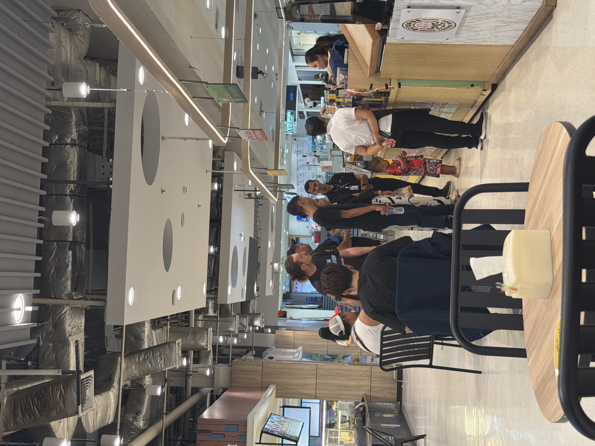 The image depicts an indoor scene, likely a cafe or food service area within a commercial establishment, with an overhead view of the ceiling showing extensive ductwork and lighting fixtures. In the foreground, a round wooden table with black metal chairs is visible, set with a dispenser of tissues.
The midground features a group of individuals in various states of interaction. On the left, several people are seated at tables and standing, appearing to be customers. A man with a distinctive ponytail is in conversation with another man wearing a black t-shirt. To their right, a woman is interacting with a male employee behind a counter. Further to the right, a young child in a colorful outfit is standing near the counter.
The background reveals more of the establishment, including more seating, counters with food or drink displays, and signage. The overall environment suggests a modern, well-lit commercial space, possibly a food court or a section of a larger store. The presence of employees in uniforms indicates a service-oriented setting. The specific location within Ban Bang Rak, Thailand, is not definitively ascertainable from the visual information alone, but the overall setting is consistent with a contemporary urban commercial area in Thailand.