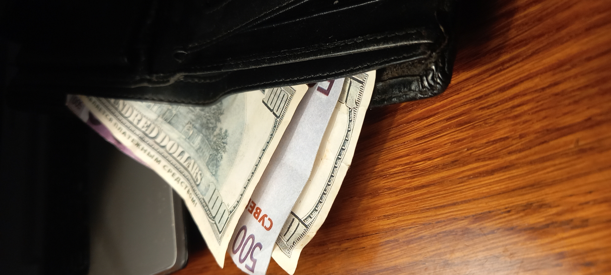 The image presents an overhead view of a black, worn leather wallet with various banknotes partially fanned out from its interior. The wallet is positioned on a polished wooden surface, which exhibits a distinct linear grain pattern.
Inside the wallet, several banknotes are visible. Prominently featured are what appear to be genuine US 100 dollar bills, displaying the text "HUNDRED DOLLARS" and the numeral "100". Among these, there are two other notes. One is a purple-colored note clearly marked with the numeral "500" and the Ukrainian text "П'ЯТСОТ ГРИВЕНЬ" (Five Hundred Hryvnia). This note also contains the Russian text "СУВЕНИРНАЯ ПРОДУКЦИЯ" (Souvenir Product), indicating its novelty status. Another lighter-colored note, partially seen, shows the numeral "100" and the Russian text "НЕ ЯВЛЯЕТСЯ ПЛАТЕЖНЫМ СРЕДСТВОМ" (Is Not A Means Of Payment), also identifying it as a souvenir or prop.
A dark, rectangular object, possibly a smartphone, is partially visible underneath the wallet and banknotes on the left side of the frame. The scene depicts money, a mix of genuine currency and souvenir notes, being stored within a wallet. The inclusion of Ukrainian and US currencies, alongside souvenir notes, is consistent with the provided location context of Kryvyi Rih, Ukraine. The lighting suggests an indoor setting.
