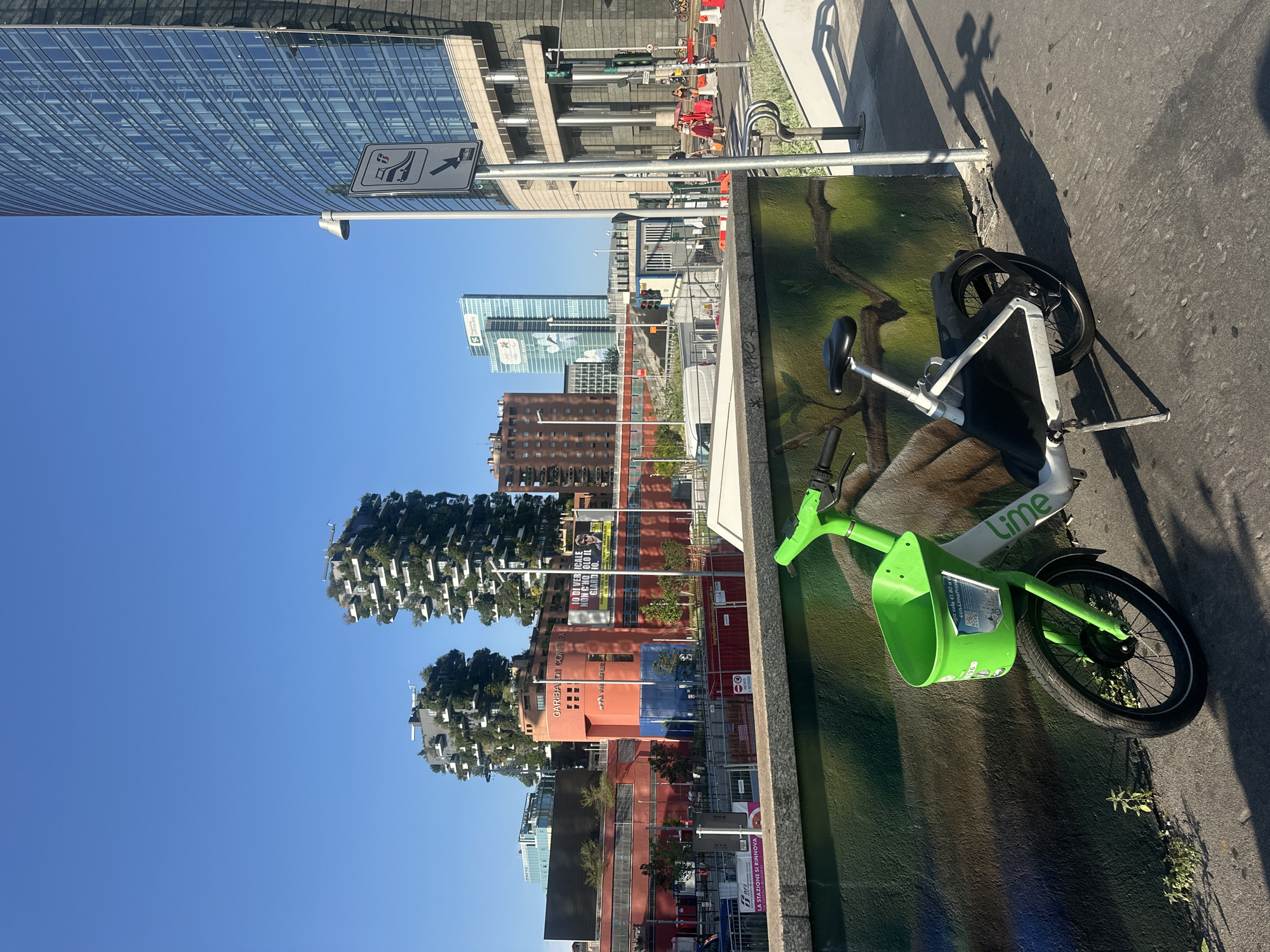 A Lime electric scooter is parked in front of a mural on a wall in Milan, Italy, on a clear, sunny day. The scooter is green and white with a lime green basket on the handlebars. Behind the scooter, a modern cityscape unfolds, featuring the iconic Bosco Verticale (Vertical Forest) residential towers, which are covered in trees and greenery. To the right, a tall, glass-faced skyscraper dominates the background. In the middle ground, there are more buildings, including a red structure with orange accents and signage, and a tall blue skyscraper in the distance. A street sign indicating a pedestrian crossing is visible next to the tall skyscraper. Several people are walking on the sidewalk in the distance, and a few more scooters are parked further down the street. Construction barriers and fencing are present in the mid-ground, suggesting ongoing development or renovation. The ground in the foreground is asphalt pavement, and the wall with the mural has a dark green background with images of branches and leaves.