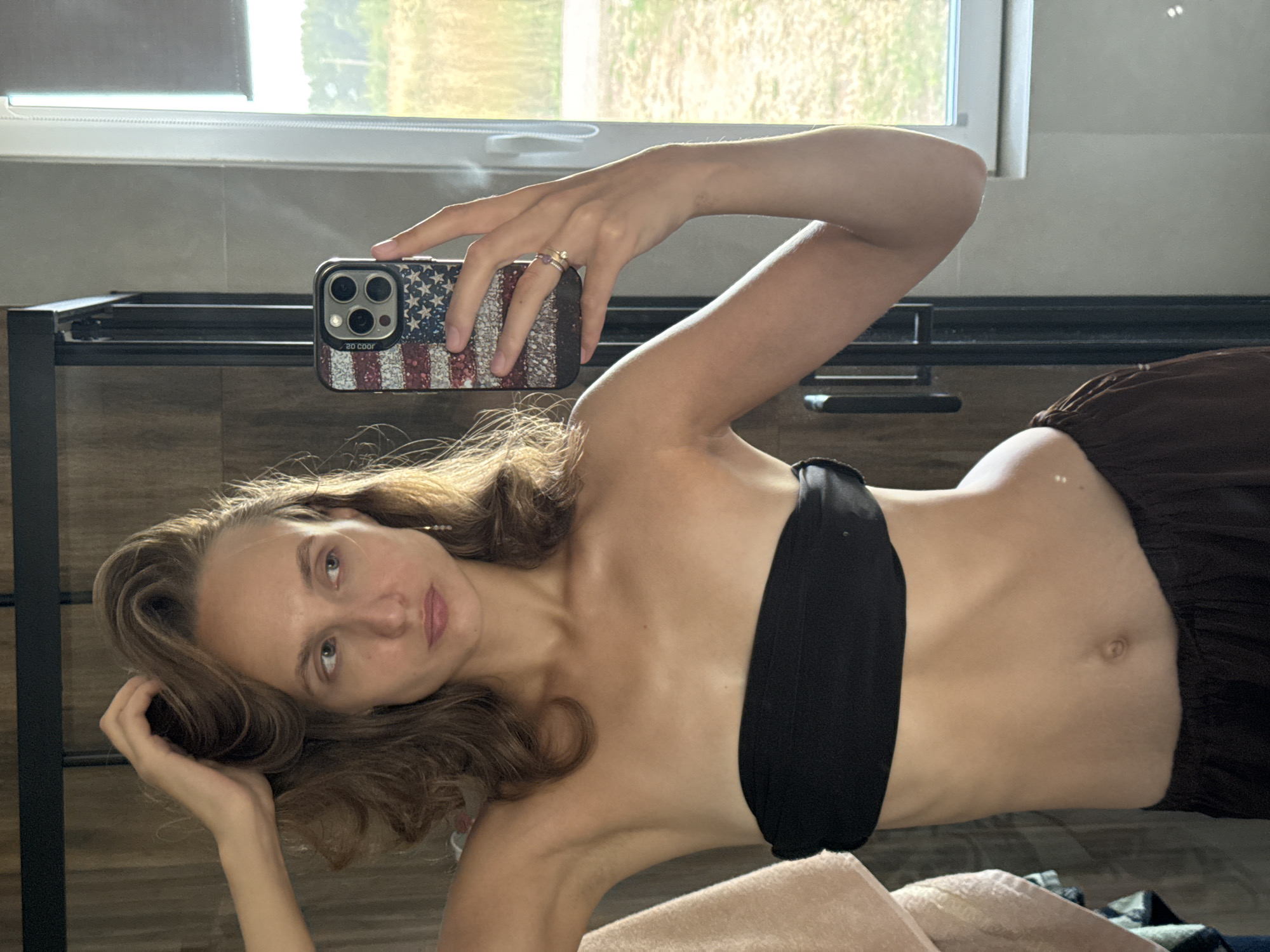 A young woman is taking a mirror selfie in a bathroom. She is wearing a black strapless top and brown pants. Her right hand is raised to her hair, and her left hand holds a smartphone with an American flag-themed case. The background shows a tiled wall, a glass shower door, and a window with a view of a rural landscape. The image is lit by natural light coming from the window.
