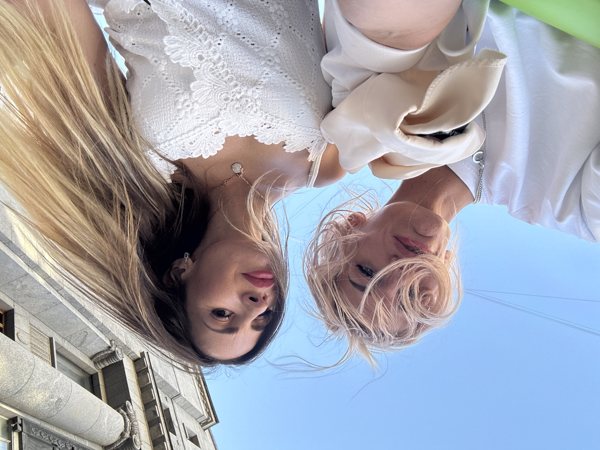 Two young women are captured in a low-angle selfie, their faces close together and looking upwards towards the camera. The woman on the left has long, dark blonde hair that cascades around her shoulders. She is wearing a white lace top and a delicate necklace with a small circular pendant. Her makeup features bright pink lipstick and dark eyeliner. The woman on the right has short, blonde hair that frames her face. She is wearing a white garment with a draped, cream-colored fabric. She has subtle makeup on, and her eyes are focused on the camera.
The background is a clear, light blue sky, suggesting daytime with good weather. The perspective of the photo, with the women appearing upside down and looking up, implies they are standing and the photo is taken from below, possibly looking up at a building. To the left of the women, a portion of a building facade is visible. It appears to be made of stone, with classical architectural elements such as columns, decorative carvings, and windows. This architectural style, combined with the context of Saint Petersburg, Russia, suggests the photo was taken in a historic district of the city. The overall mood is casual and friendly, with the women appearing relaxed and happy. There is no visible text in the image.