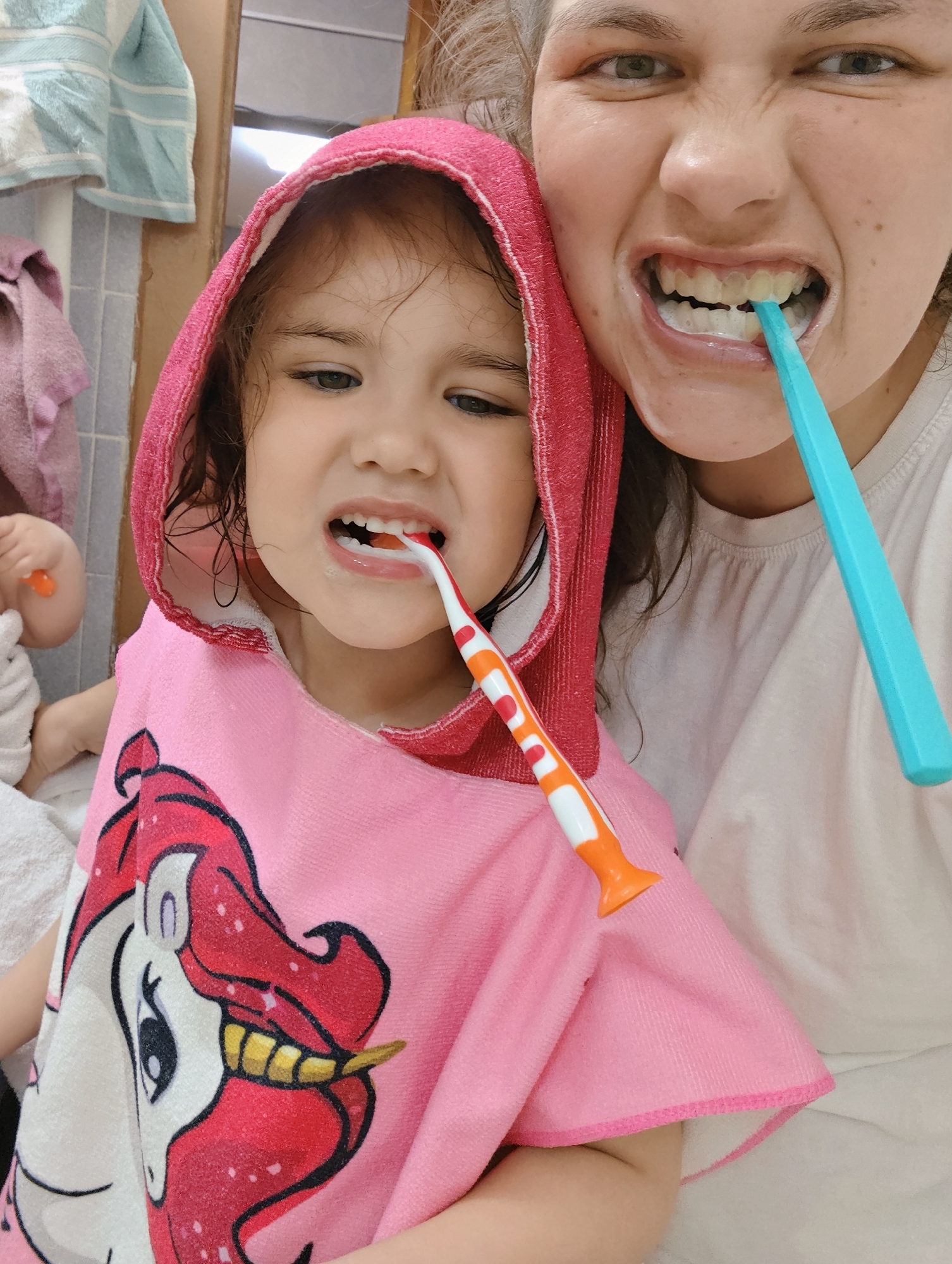 The image shows a young girl and a woman in a bathroom, both brushing their teeth. The girl is wearing a pink hooded towel with a unicorn design and is holding an orange and white toothbrush. The woman is holding a blue toothbrush and shows her teeth. The bathroom contains blue and white towels. A third person with an orange toothbrush is partially visible behind the girl. The location is in the city of Chaikovsky, Russia.