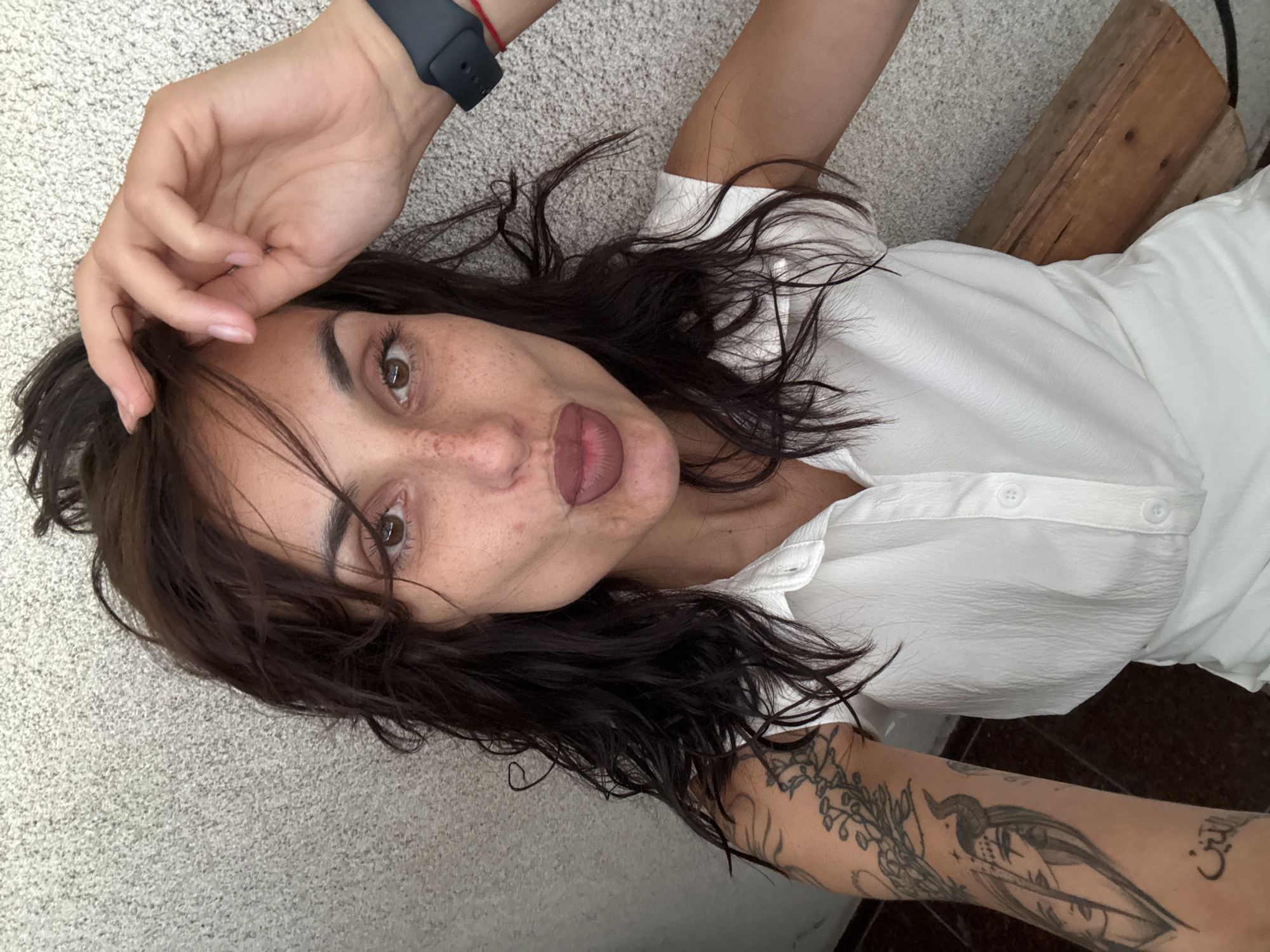A young adult female with dark, messy hair, freckles, and full lips puckered is taking a selfie. She is wearing a white collared shirt and has tattoos on her left arm, including a stylized face with horns. She has a smartwatch on her right wrist. The background appears to be a textured wall and a wooden bench. The scene is likely an outdoor urban setting in Pleven, Bulgaria, possibly a public park or a residential area.
