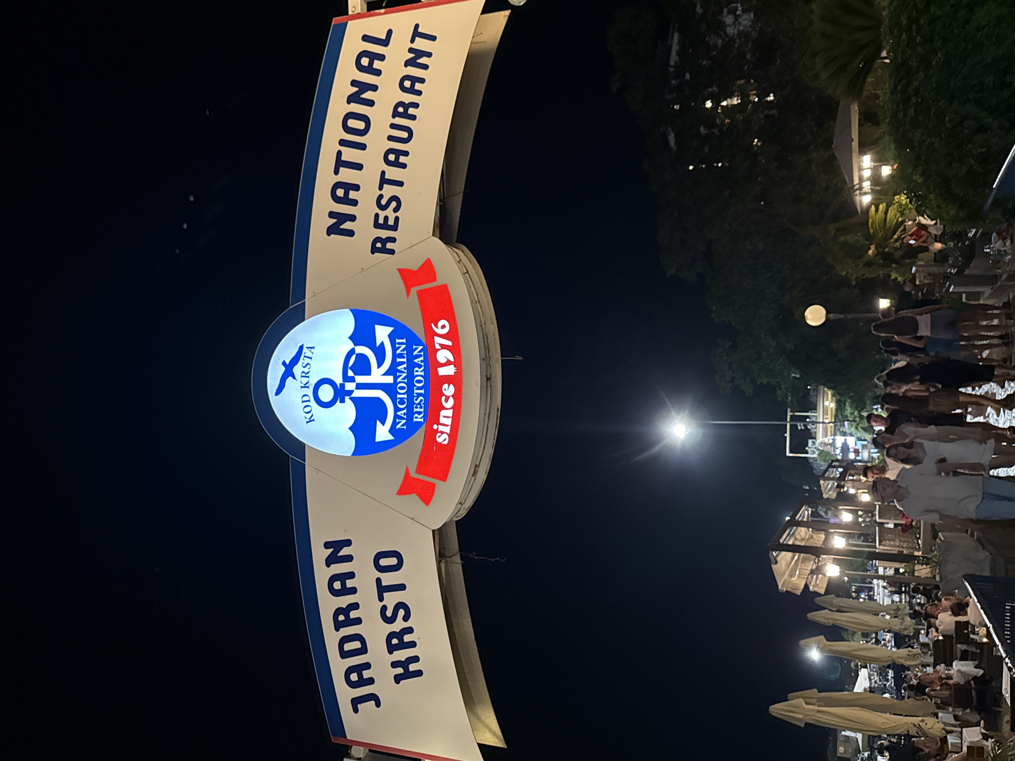 The image shows a nighttime scene in Budva, Montenegro, featuring the illuminated sign of "JADRAN KRSTO NATIONAL RESTAURANT" with the founding year "since 1976". The sign is positioned above an outdoor dining area where people are seated at tables under umbrellas, illuminated by various lights. Pedestrians are visible walking along a pathway in front of the restaurant. The background consists of trees and buildings, suggesting a coastal or waterfront location. The sky is dark, indicating it is nighttime, and a few lights are visible in the distance.