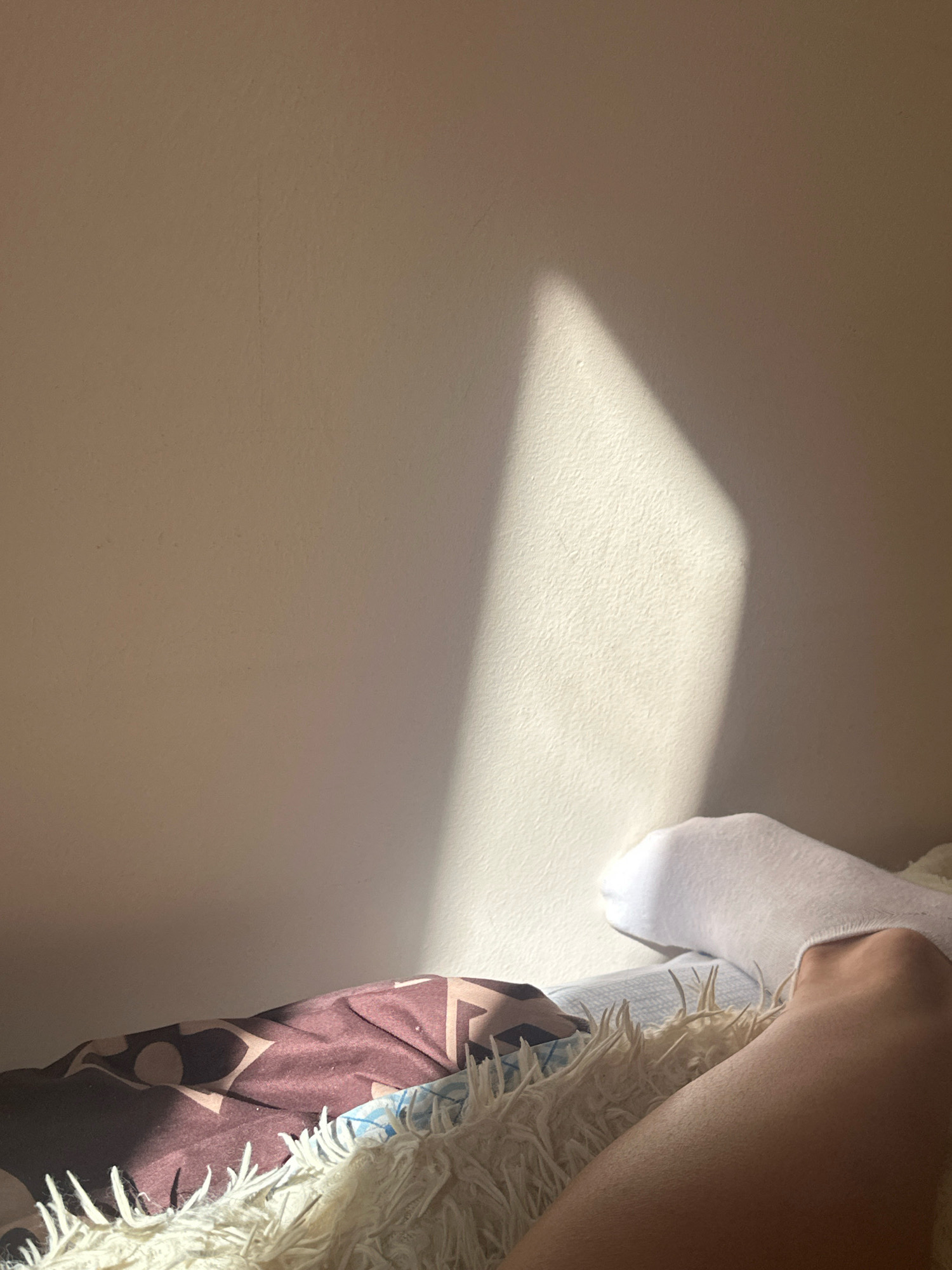 A close-up, eye-level shot captures a portion of a person's legs and feet resting on a bed, with a wall in the background. The person is wearing a white sock on their right foot, and their bare left leg is visible, extending towards the foreground. The bed is covered with a patterned blanket and a fluffy white throw. A diagonal beam of sunlight cuts across the wall, casting a bright, sharp shadow. The overall scene is domestic, suggesting a moment of relaxation in a bedroom. The image does not contain any information that would allow for the specific identification of the location as Gyumri, Armenia.