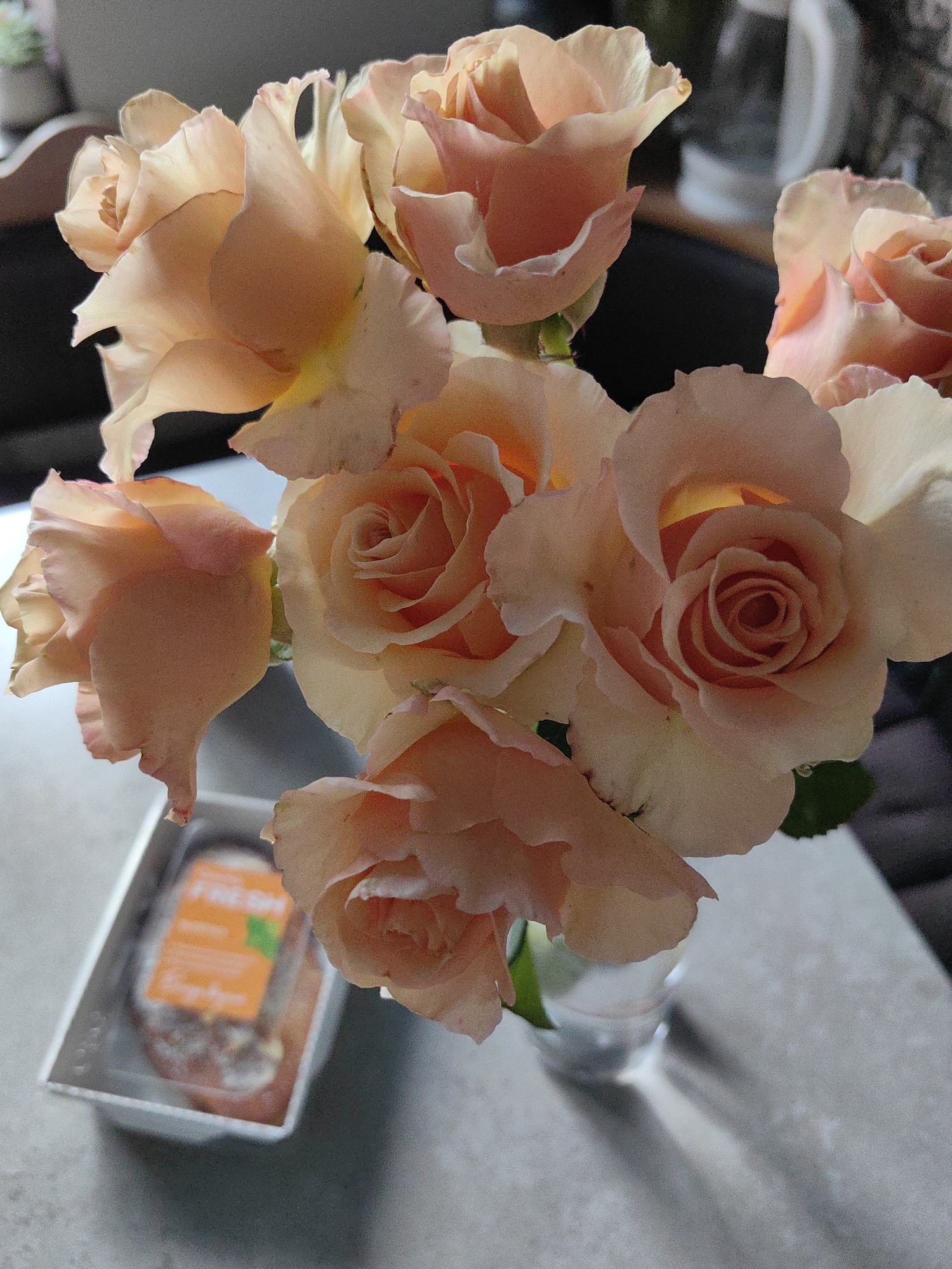 In Fanipal, Belarus, a close-up shot captures a bouquet of peach-colored roses in a clear glass vase, resting on a textured, light-colored surface, possibly a table. The lighting creates dramatic shadows on the surface, highlighting the delicate petals of the roses. To the left of the vase, a white plastic container with a clear plastic lid holds a product with an orange label displaying the word "FRESH" in white letters. The background is blurred, suggesting an indoor setting. There are no people visible in the image.