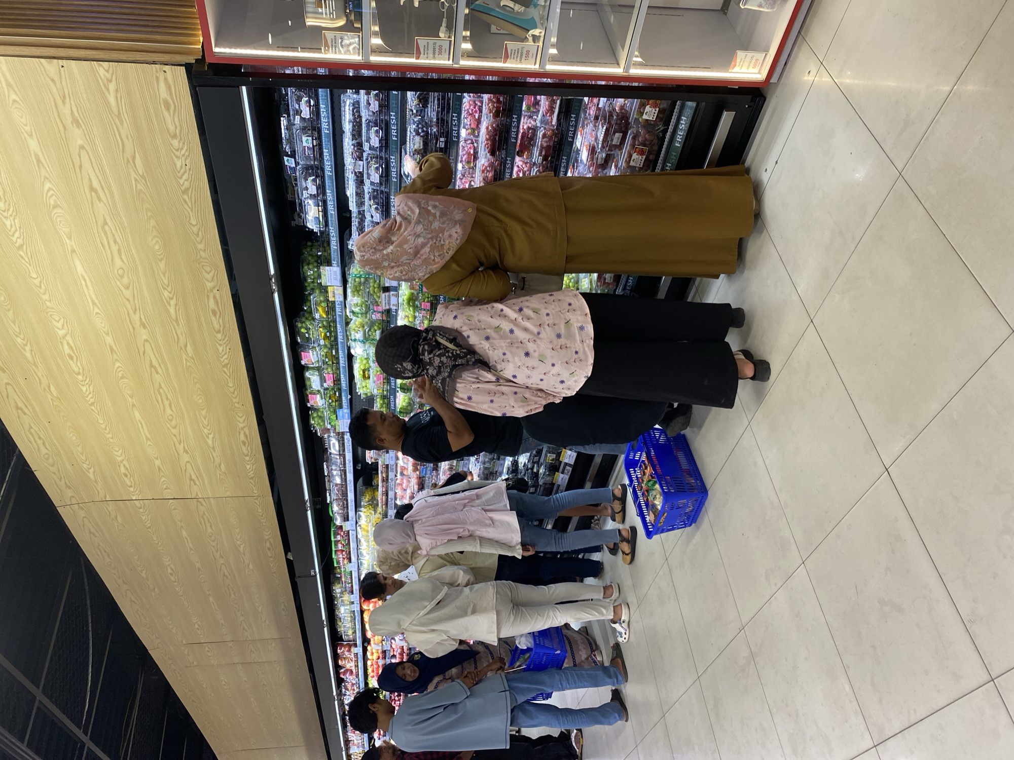 The image depicts a brightly lit interior of a modern supermarket in Banda Aceh, Indonesia, specifically focusing on a produce section with refrigerated display cases. Several individuals, predominantly women in modest Islamic attire, are seen browsing and selecting fruits from the shelves. The display cases are filled with various types of fruits, including grapes and what appear to be berries, neatly packaged in transparent containers. The floor is tiled with large, light-colored squares. A blue plastic shopping basket filled with items is visible on the floor in the foreground. The overall atmosphere suggests a routine shopping experience in a well-stocked grocery store.