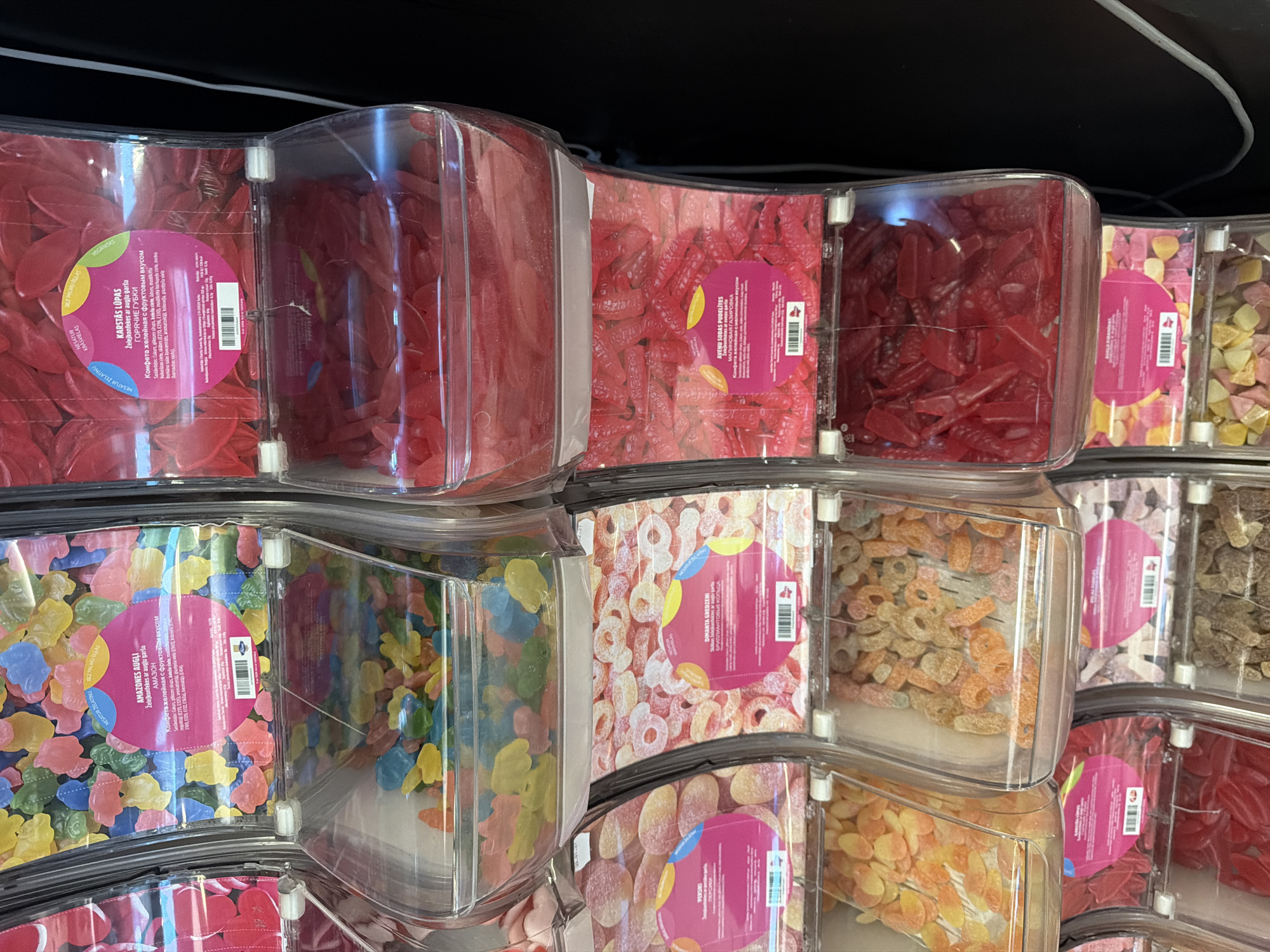 The image depicts a detailed view of a multi-tiered bulk candy display in a retail setting, likely a supermarket or confectionery store in Riga, Latvia. The display consists of clear plastic gravity dispensers, arranged in vertical columns and horizontal rows, filled with various types of colorful gummy and jelly candies.
In the top left dispenser, multi-colored, irregular blob-shaped gummy candies, labeled "AMAZONES AUGLI" (Amazon Fruits) in Latvian and "АМАЗОН" in Russian, are visible. To its right, red, flat, leaf-shaped gummy candies are displayed, labeled "KARSTĀS LŪPAS" (Hot Lips) in Latvian and "ГОРЯЧИЕ ГУБЫ" in Russian.
Below these, on the left, are peach-colored, oval-shaped jelly candies identified as "PERSIKI" (Peaches) in Latvian and "ПЕРСИКИ" in Russian. Adjacent to them, in another dispenser, are sugar-dusted, peach-colored ring-shaped candies, labeled "DZĪMĀKTA GREDZENI" (Chewy Rings) in Latvian and "ЖЕВАТЕЛЬНЫЕ КОЛЬЦА" in Russian.
Further down, red, small bottle-shaped gummy candies are labeled "AYENU SODAS PUDELĪTES" (Raspberry Soda Bottles) in Latvian and "МАЛИНОВАЯ ГАЗИРОВКА" (Raspberry Soda) in Russian. To their right, red, chili pepper-shaped gummy candies are present, labeled "Ābolu pipariņi" (Apple Peppers) in Latvian and "Яблочные перцы" in Russian. Other dispensers show yellow/orange flat candies and darker, possibly brown, irregularly shaped candies.
Each candy type has a distinct pink circular label adhered to the dispenser, featuring product names, descriptions, and other information in both Latvian and Russian languages, alongside a barcode. The consistent bilingual labeling in Latvian and Russian confirms the location within Latvia. The background is a dark, uniform surface, with a white electrical cable running vertically on the far right. The scene is brightly lit, emphasizing the vibrant colors and textures of the candies. No individuals are visible in the frame.