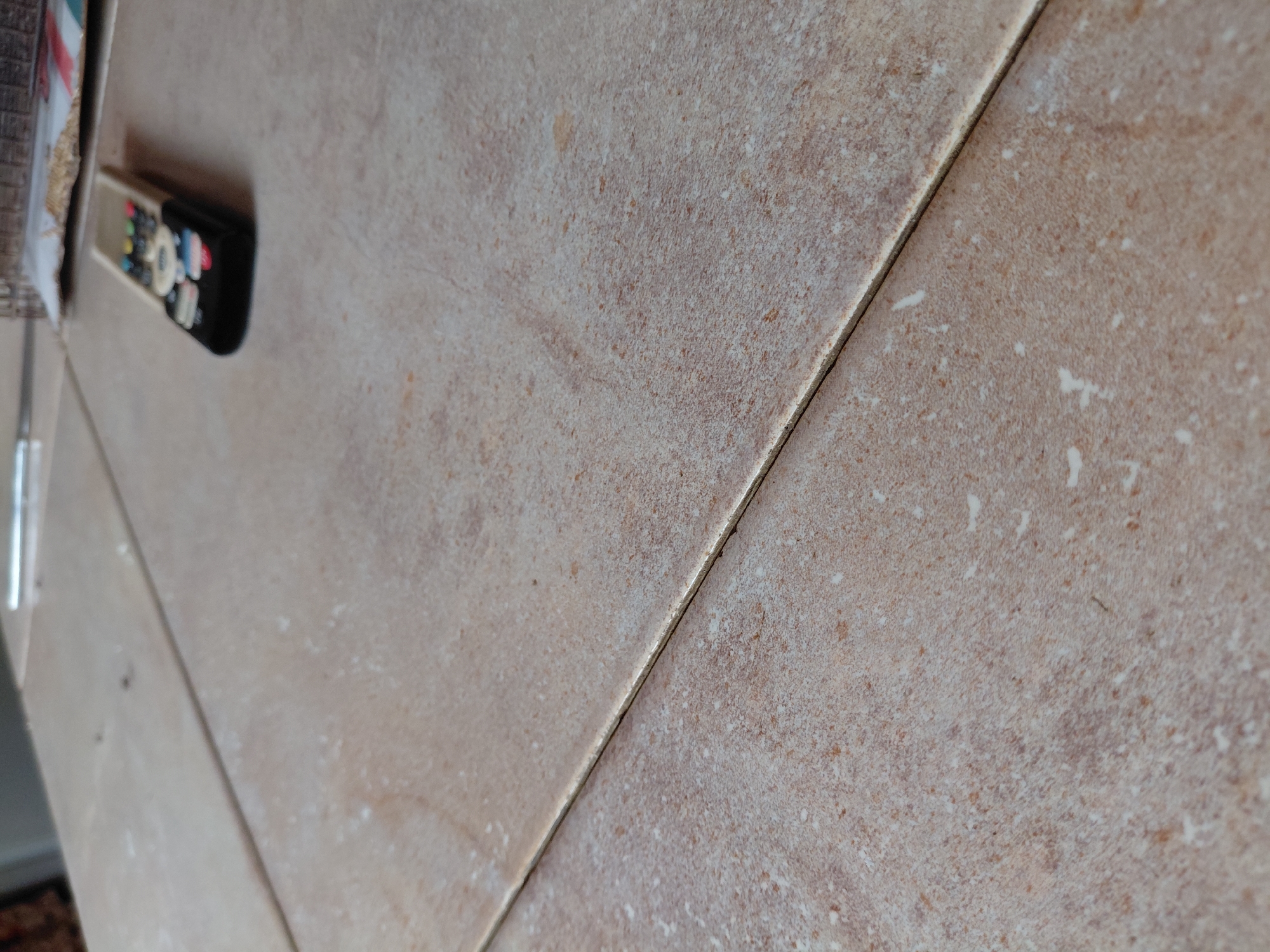 The image presents an angled, close-up perspective of a light brown or beige tiled surface, likely a floor or countertop, in an indoor setting. The tiles exhibit a speckled, textured pattern with variations in light and dark brown hues, and subtle, lighter streaks. A prominent dark grout line diagonally separates two large tiles, running from the top right towards the bottom left of the frame.
On the left side of the tiled surface, a remote control is partially visible. It has a light-colored body with a black top and several distinctively colored buttons, including red, blue, and yellow, though no text is legible on them. Above the remote, a fragment of a white and red patterned object, possibly a book or magazine, can be seen. Below the remote, in the bottom-left corner, a sliver of a darker, possibly patterned floor covering, such as a rug, is visible. A thin, reflective, silver-colored object, resembling a pen or a rod, is barely discernable along the far left edge, extending upwards.
The scene depicts a static arrangement of common household items. No individuals are present, and no specific activity is taking place. The lighting suggests an interior environment, but does not provide clues about the time of day or outdoor weather. The image itself offers no explicit visual cues to specifically identify the location as Santa Ana, Venezuela, apart from the provided context.