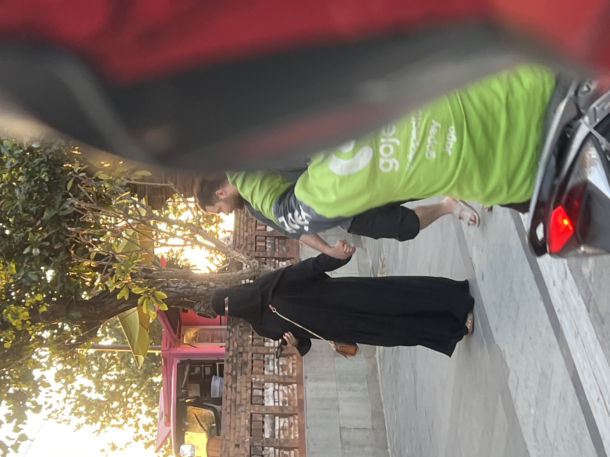 Two individuals are walking hand-in-hand on a paved pedestrian pathway in Kuta, Indonesia. The person on the left is wearing a full-length black niqab and a brown crossbody bag. The person on the right is wearing a bright green shirt with "goje" and other text in white lettering, and black shorts. The background shows a brick wall, a pink food truck with an umbrella, and trees with sunlight filtering through the leaves. The foreground is partially obscured by the rider's helmet and motorcycle.