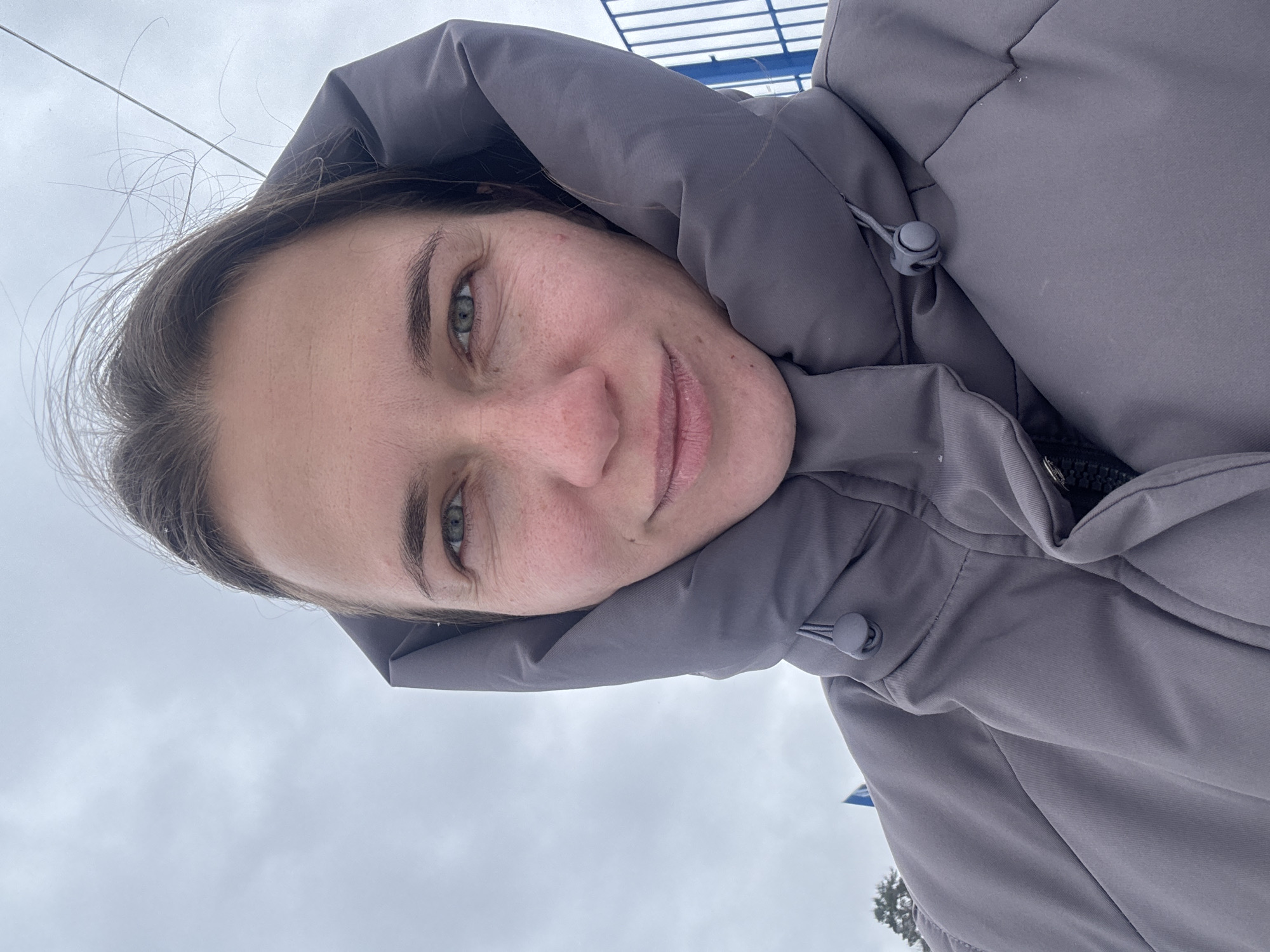 The image is a close-up, slightly tilted portrait of a woman taking a selfie outdoors in Раздолье, Russia. She is wearing a practical, grey hooded winter jacket, with the hood pulled up around her head. Her head is tilted, and she looks directly at the camera with a gentle, relaxed expression and a subtle smile.
Notable details include her green eyes and visible freckles on her nose and cheeks. Her dark hair is partially visible, with some loose strands appearing windswept, suggesting a breezy outdoor environment. The jacket features a drawstring with a grey toggle on the hood.
The background is predominantly an overcast, light grey sky, indicating a cloudy daytime. Faint blue structural elements, possibly part of a metal framework like a ski lift or outdoor facility, are visible in the upper background. A subtle hint of a tree can also be discerned in the lower background. The attire and sky condition suggest cold or chilly weather. There is no visible text in the image.
