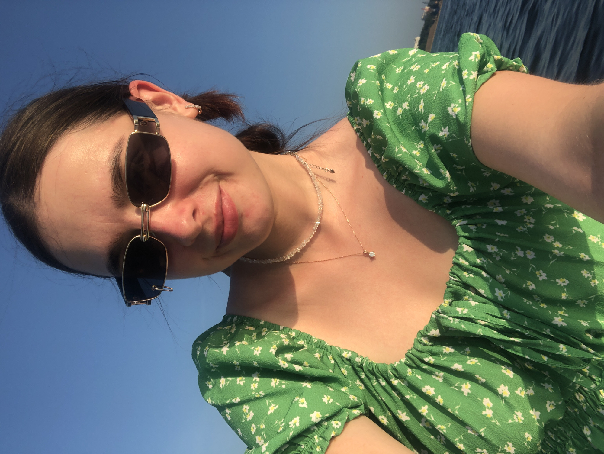 A young woman with dark hair pulled back into a ponytail and wearing a green floral dress and dark sunglasses is captured in a selfie. She is adorned with a beaded necklace and a pendant. Her left ear is visible, featuring a small hoop earring. The background consists of a clear blue sky and dark blue water, suggesting an outdoor setting, possibly near the Kama River in Chaikovsky, Russia, given the presence of a distant shoreline and buildings in the upper right portion of the frame. The lighting indicates it is late afternoon or early evening, with the sun casting a warm glow on her skin.