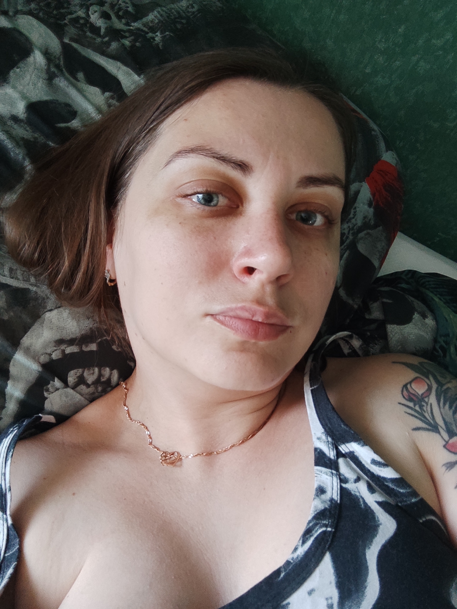 A young adult Caucasian woman with medium-length brown hair and blue eyes is shown in a close-up, overhead shot. She is wearing a black and white patterned tank top and a rose gold necklace with a heart pendant. A tattoo of a flower is visible on her left shoulder. She appears to be lying down, possibly in a bed, with patterned sheets in the background. The lighting is natural and appears to be coming from above. The background is out of focus, suggesting shallow depth of field. The woman is looking directly at the camera with a neutral expression. There are no other people or objects of note in the frame.