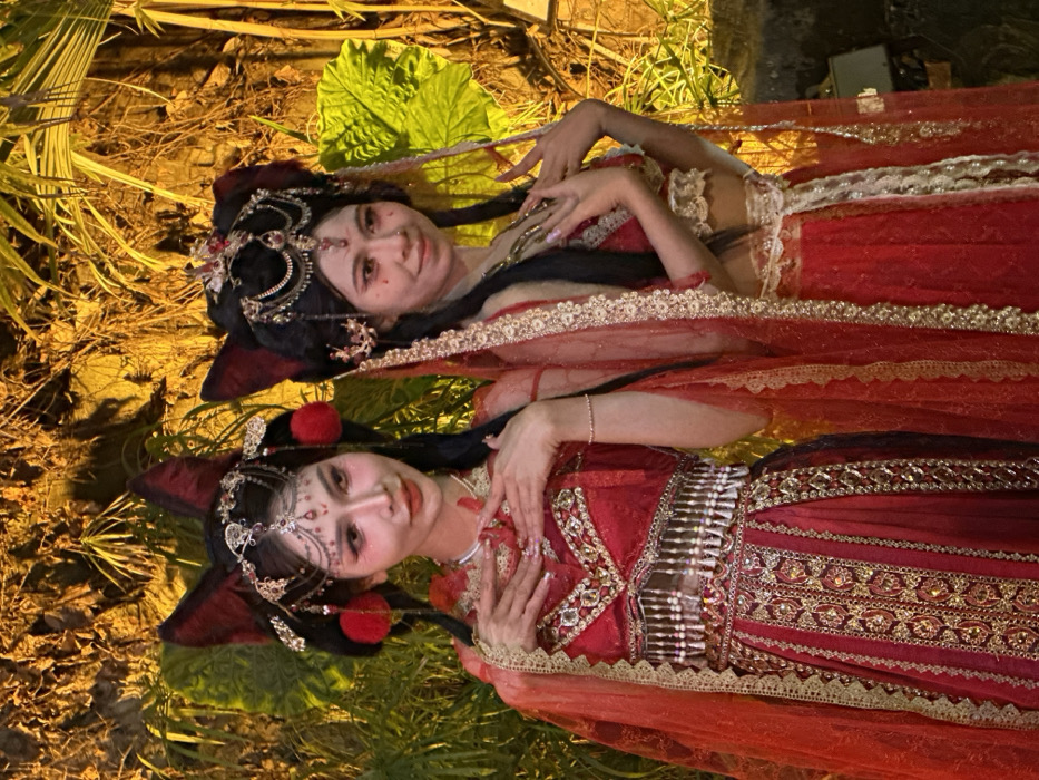 The image displays two East Asian females posing outdoors in Yuzhong District, China. Both individuals are dressed in elaborate, historically-inspired red and gold costumes featuring intricate embroidery and embellishments. Their hairstyles incorporate pulled-back dark hair adorned with prominent, dark red pointed ears (reminiscent of animal ears) and ornate gold-and-red jeweled headpieces that include chains draped across the forehead and red pom-poms. Both wear intricate makeup with pink and red tones around the eyes, winged eyeliner, and contoured facial features.
The individual on the left wears a two-piece costume consisting of a fitted red and gold embellished bodice with a V-neckline and a matching floor-length skirt featuring vertical gold embroidered panels and a beaded fringe at the waist. A sheer, red, gold-trimmed veil is draped over her arms and shoulders. Her hands are raised and crossed at the wrists in front of her chest, with fingers slightly splayed and manicured with long extensions. She exhibits a subtle smile, gazing slightly to her right.
The individual on the right, visible from the chest up, wears a similarly styled red and gold embellished top, possibly a bralette, with a matching skirt, and a sheer, red, gold-trimmed veil over her arms. Her hands are also raised in a similar posed manner in front of her chest. She displays a subtle smile, gazing slightly to her left.
The background consists of dense foliage, including large leaves and possibly vines, illuminated by warm, yellowish-orange artificial lighting. This suggests an outdoor, possibly landscaped, evening setting. No other architectural elements or objects are discernible in the immediate background.