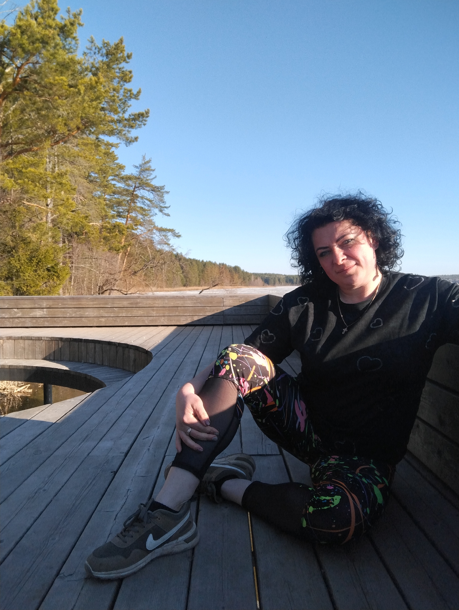 A woman is sitting on a wooden deck in an outdoor setting. She is wearing a black long-sleeved shirt with heart designs and colorful leggings. Her hair is dark and curly. She is also wearing sneakers. The deck is made of wooden planks and has a curved design. In the background, there is a frozen lake and trees. The sky is clear and blue.