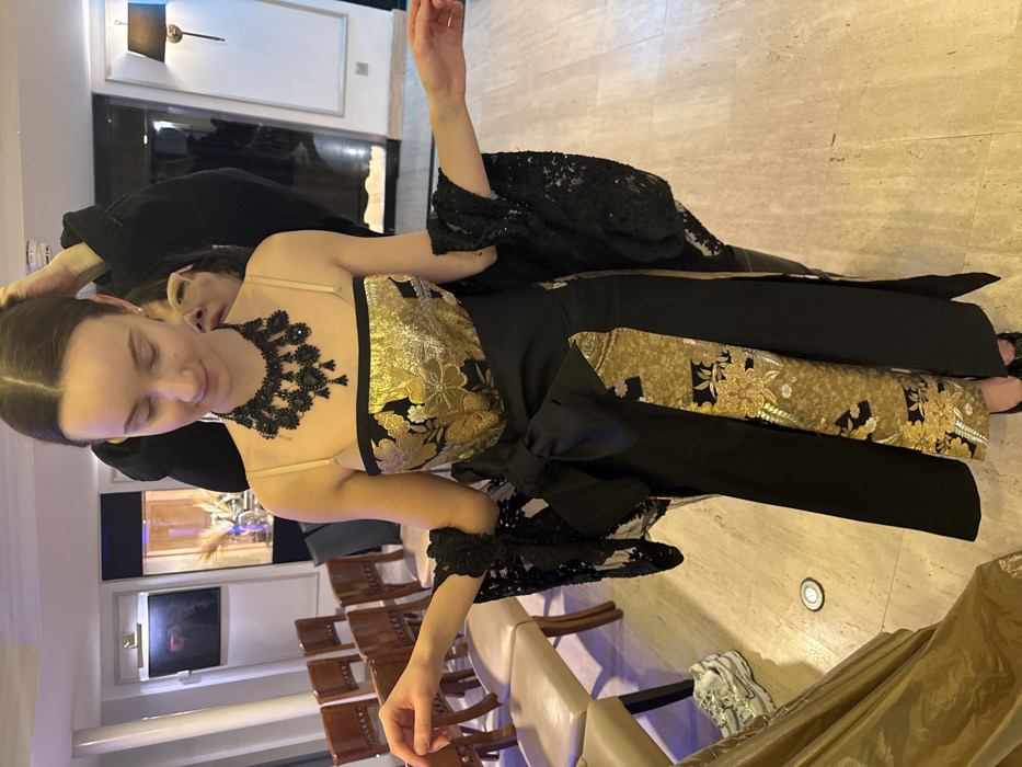 An indoor scene in an upscale venue in Paris, France, shows a young woman in the foreground being prepared for an event. She wears a strapless, floor-length gown featuring a black bodice with a wide gold and black floral patterned band, a black skirt with similar gold and black patterned vertical panels, and a large black obi-style bow at the waist. She has a prominent black beaded necklace and black lace arm coverings. Her dark hair is styled up. Behind her, partially obscured, another person is visible, with their hands near the main subject's head, appearing to adjust her hair or headwear. The main subject's arms are slightly outstretched.
The environment features light-colored, polished tile flooring. In the background to the left, several empty wooden chairs with light-colored upholstered seats are arranged in rows. Further back, a framed picture or mirror hangs on a light-colored wall with decorative molding. A display cabinet containing decorative botanicals is also visible. To the right, a wall sconce with a black lampshade is mounted on a light-colored wall. Recessed ceiling lights and a floor spotlight contribute to the illumination. On the floor to the lower left, a pair of white Nike sneakers rests beside draped golden fabric.