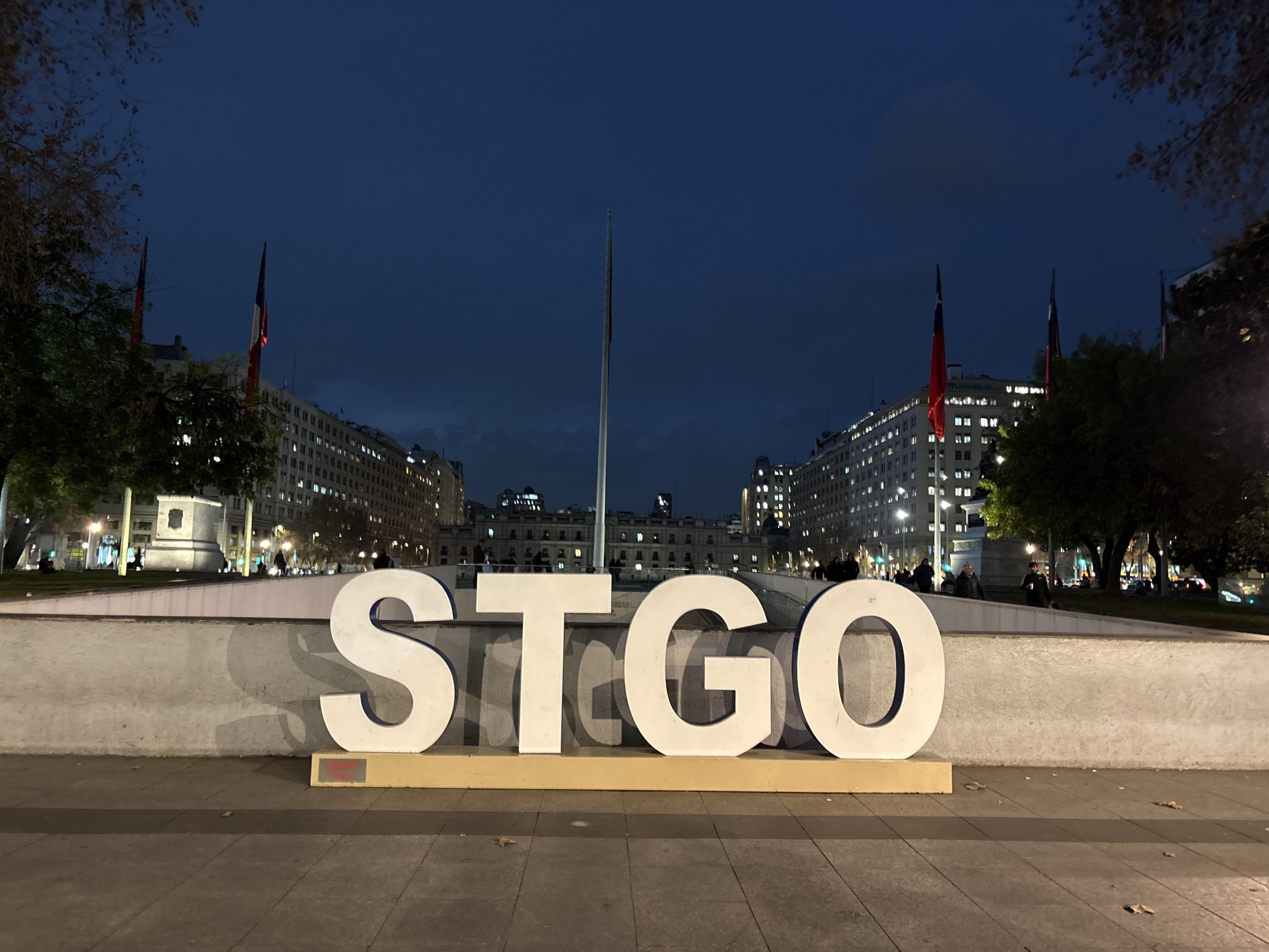 The image depicts a large, white, three-dimensional sculpture spelling out "STGO" in front of a row of illuminated buildings at dusk. The sculpture is positioned on a paved plaza, with a low, light-colored wall behind it. The background features several flags on tall poles, appearing to be Chilean flags, and tall buildings with many windows lit up. Trees with sparse foliage are visible on the left and right sides of the frame. People are sparsely scattered in the background, walking on the plaza and near the buildings. The sky is a deep blue, indicating the transition from day to night, with some subtle cloud formations. The overall scene suggests an urban setting in Santiago, Chile, likely a public square or monument area.