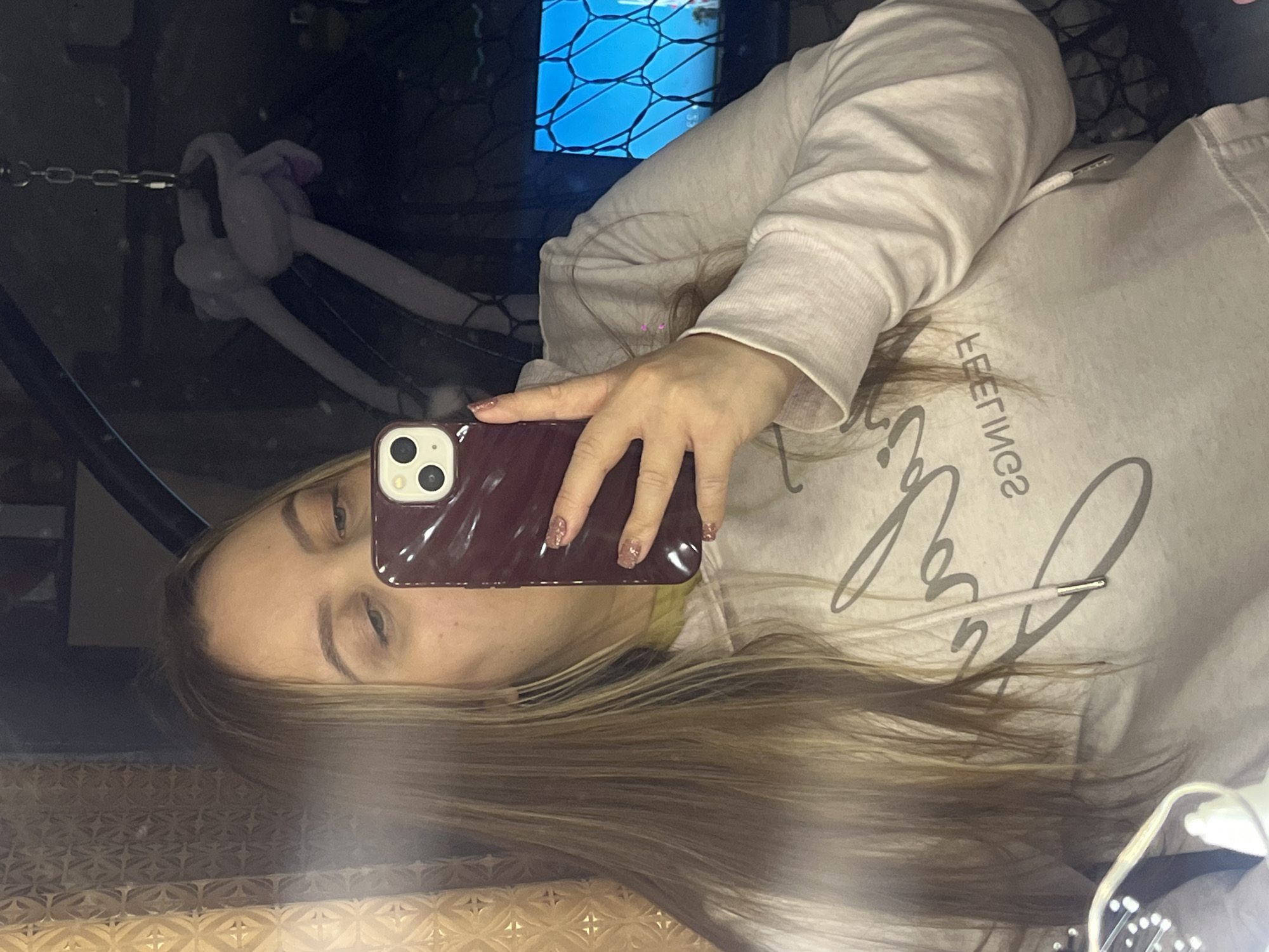 A woman with blonde hair is taking a selfie in a mirror. She is holding a maroon iPhone with a double-lens camera in her right hand, partially obscuring her face. She is wearing a light beige hooded sweatshirt with a drawstring and text printed on the front. The background is dark and appears to be inside a room. A chain and what seems to be a stuffed animal are visible in the upper left corner. A screen with a blue geometric pattern is visible behind her. The reflection in the mirror shows a gold-colored patterned surface below. There is no information to indicate that this image was taken in Novopokrovskaya, Russia.