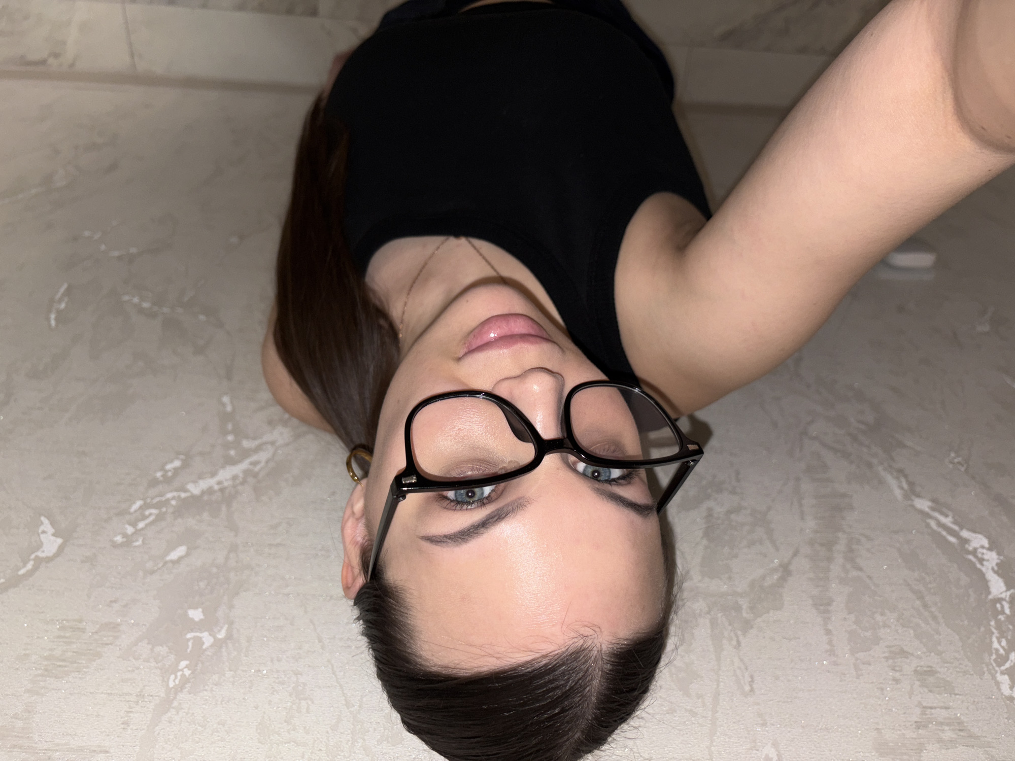 A woman with light skin and dark brown hair is lying on a light gray, speckled floor. She is wearing black-rimmed glasses and a black tank top. Her right arm is extended upwards towards the top right corner of the image. A small white object is visible near her arm. A gold necklace is visible around her neck and a small gold earring is present.
The location is not identifiable as Chaikovsky, Russia.