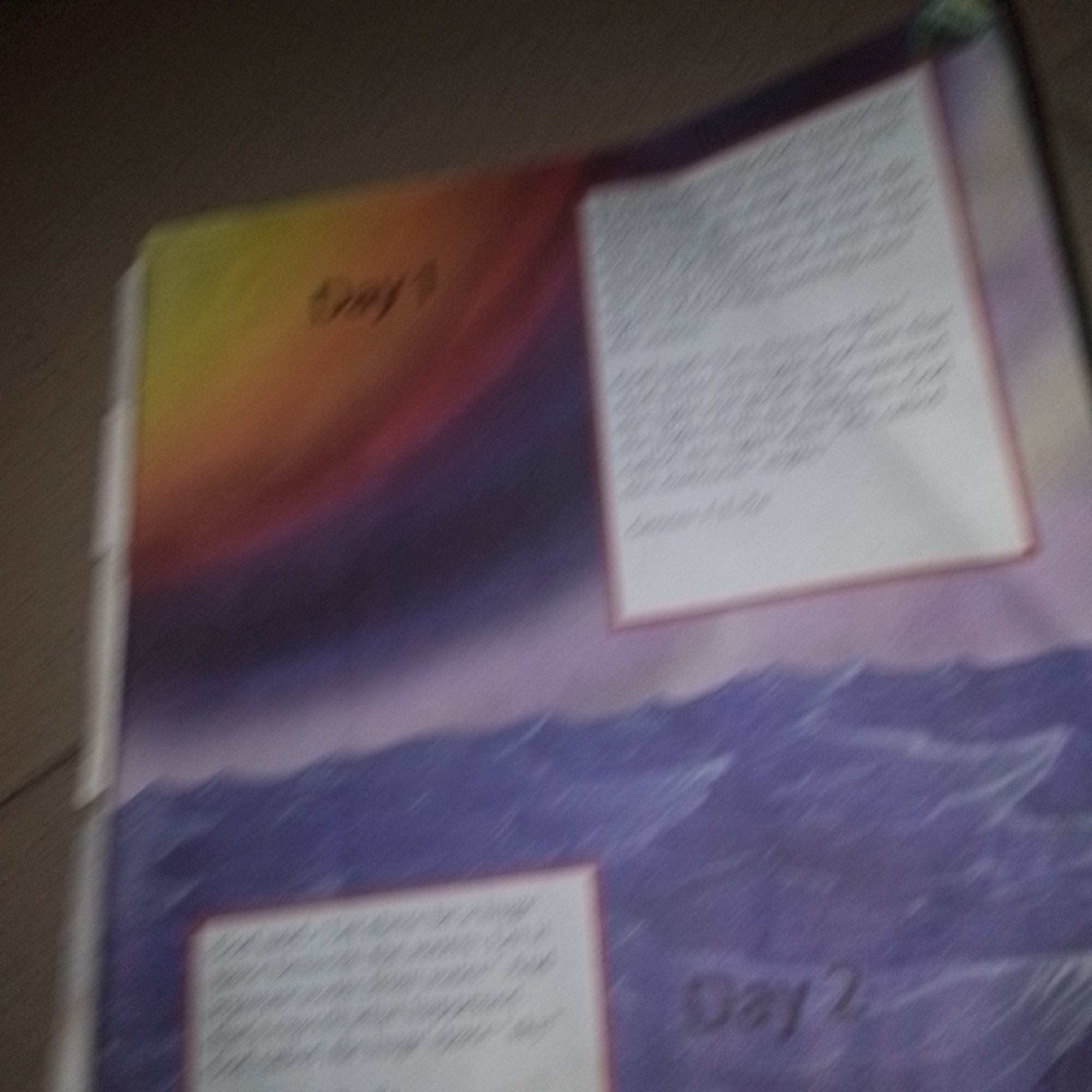 An out-of-focus, close-up shot of an open book or binder displays a colorful illustration of a sunset or sunrise over a body of water. The upper portion of the spread features a gradient of yellow, orange, and purple, depicting the sky. To the right of the sky, there is a white rectangular panel filled with text, though it is too blurred to read. Below the sky illustration, the image transitions to a purple and blue depiction of water with white lines suggesting waves. On the lower right side of the spread, another white rectangular panel with text is visible, also illegible due to blurriness. To the left of this lower text panel, the words "Day 2" are written in dark, smudged lettering. The overall image quality is poor, with significant motion blur and lack of focus, making specific details and context impossible to ascertain. The scene appears to be an indoor environment, possibly a table or floor, given the dimly lit background.