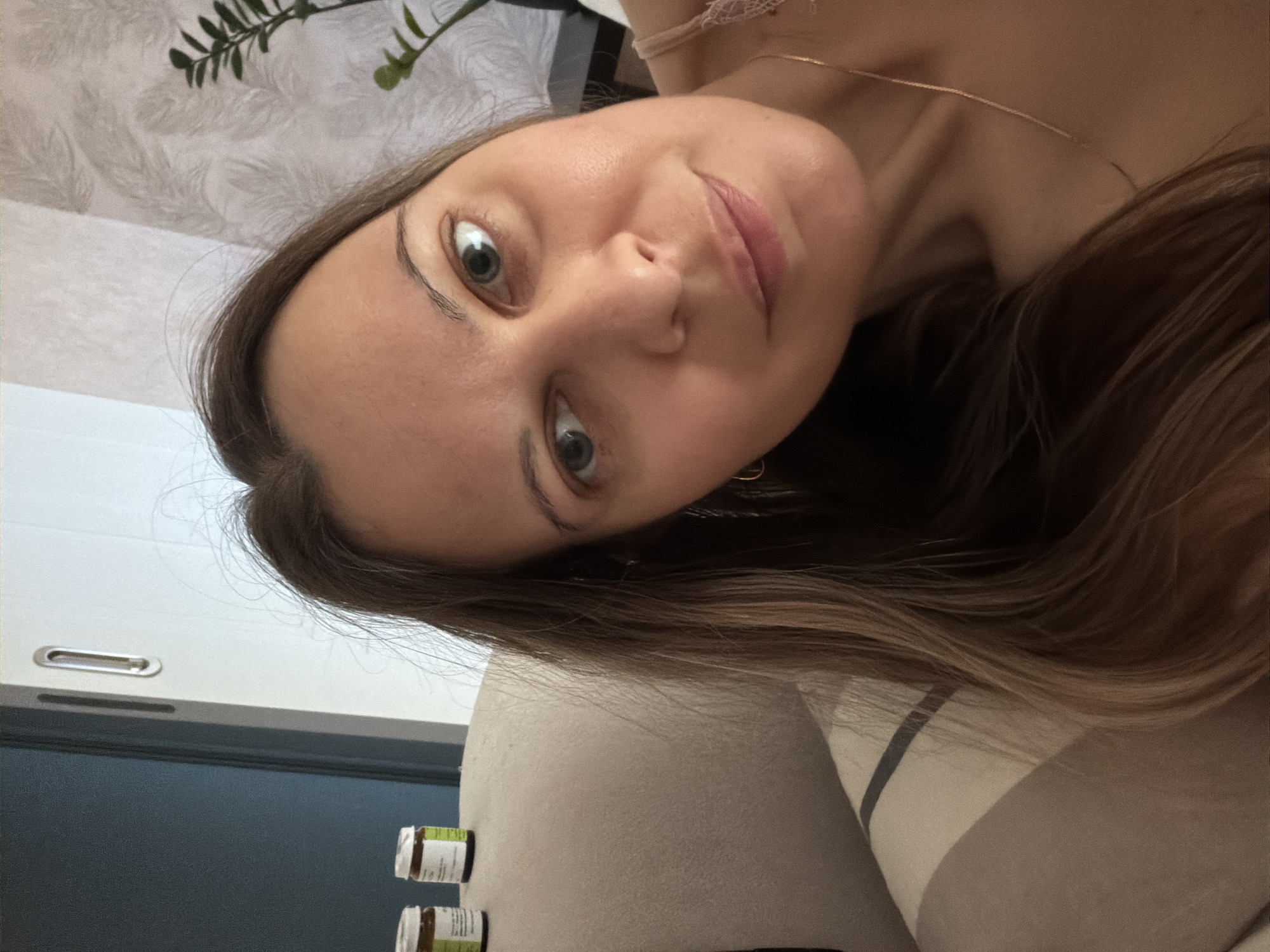 The image is a selfie taken indoors. A woman with long brown hair and light eyes is the primary subject. She is looking towards the camera with a slight smile. She is wearing a light-colored camisole with lace trim and a delicate gold necklace. Her face is illuminated by soft light, highlighting her skin. In the background, on what appears to be a beige couch, are two small brown jars with white labels. To the left, a dark blue door is partially visible, and to the right, a wall with a textured, light-colored wallpaper is in the background, with a green plant and a lamp. The overall setting suggests a domestic environment, possibly a living room or bedroom. There are no clear indicators to suggest the location is specifically in the city of Izhevsk, Russia.