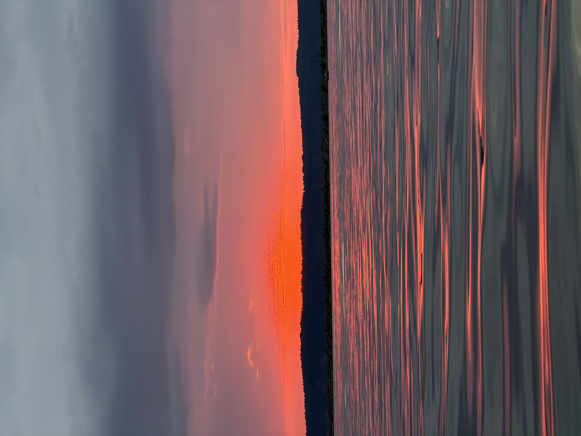 A serene sunset over the Kama River in Chaikovsky, Russia. The sky is a vibrant gradient of deep gray clouds at the top, transitioning to soft pinks and fiery oranges as it nears the horizon. A silhouette of a tree-lined hill forms the backdrop, with a few faint structures visible on the distant shore. The foreground is dominated by the calm surface of the Kama River, reflecting the colors of the sunset in long, horizontal streaks of orange and gray. The water appears smooth with gentle ripples, creating an almost abstract pattern of light and shadow. A small boat is visible on the right side of the river, suggesting human presence, though no individuals are clearly discernible. The overall atmosphere is peaceful and tranquil.