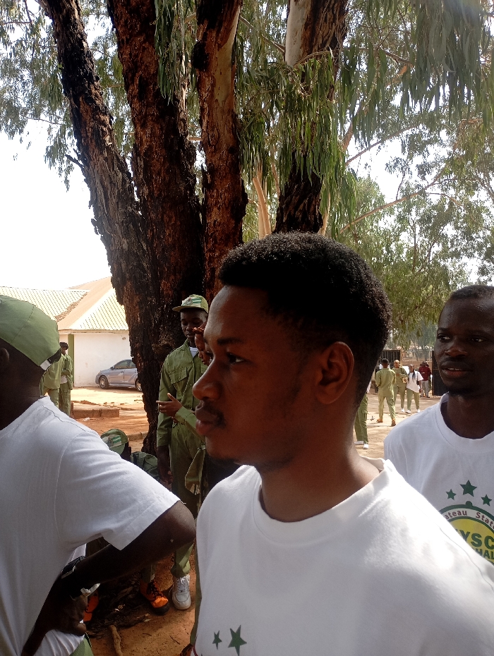 The image captures a group of people in Uke, Nigeria, near a building with a white roof. The scene is outdoors, beneath the shade of several tall trees. The central foreground features a man in a white t-shirt with a dark green star, facing right. To his right and slightly behind him stands another man in a similar white t-shirt. Behind the central figure are multiple people in green military uniforms, suggesting a gathering or event related to the military. A car is parked behind the building. The ground is a reddish-brown dirt.