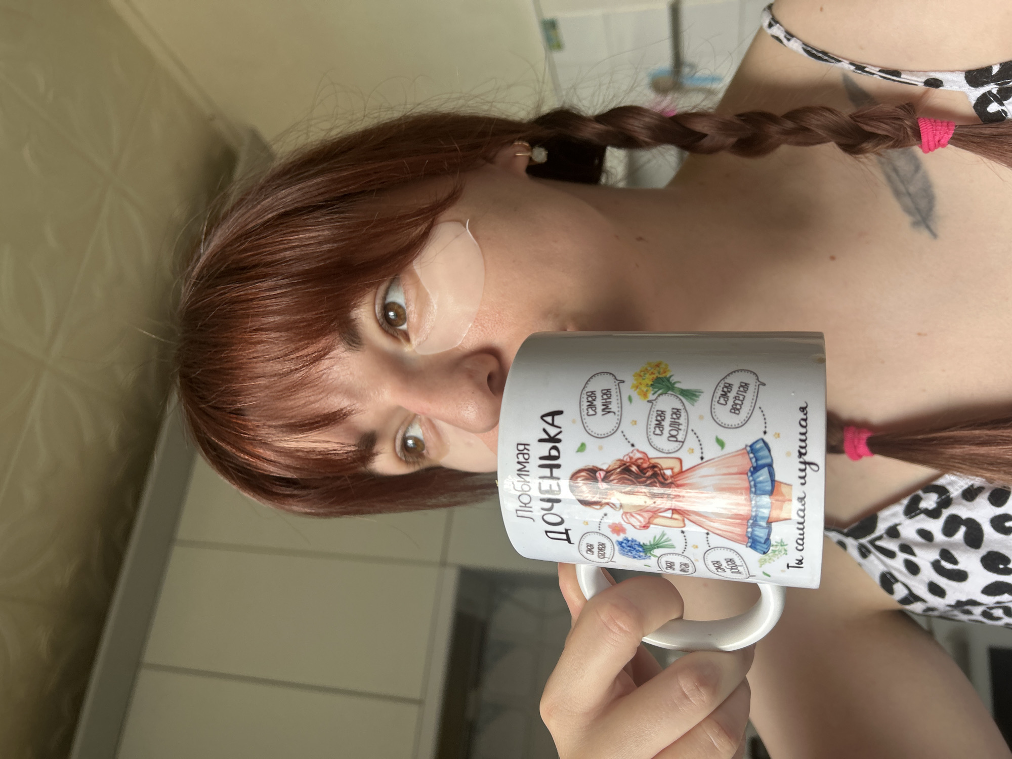 A woman with reddish-brown hair, styled in braids with pink hair ties, holds a white mug with an image of a girl in a dress, flowers, and text in Russian. The woman has an under-eye patch on her left eye and is positioned in front of a light-colored wall with visible seams. A small portion of a black and white patterned garment is visible on her shoulder. The text on the mug reads "Любимая ДОЧЕНЬКА" (Favorite Daughter), "самая умная" (smartest), "самая родная" (most native), "самая красивая" (the most beautiful), "самая Весёлая" (the most cheerful), "Ты самая лучшая" (you are the best). The precise location, Фоки, Russia, is not evident from the image.