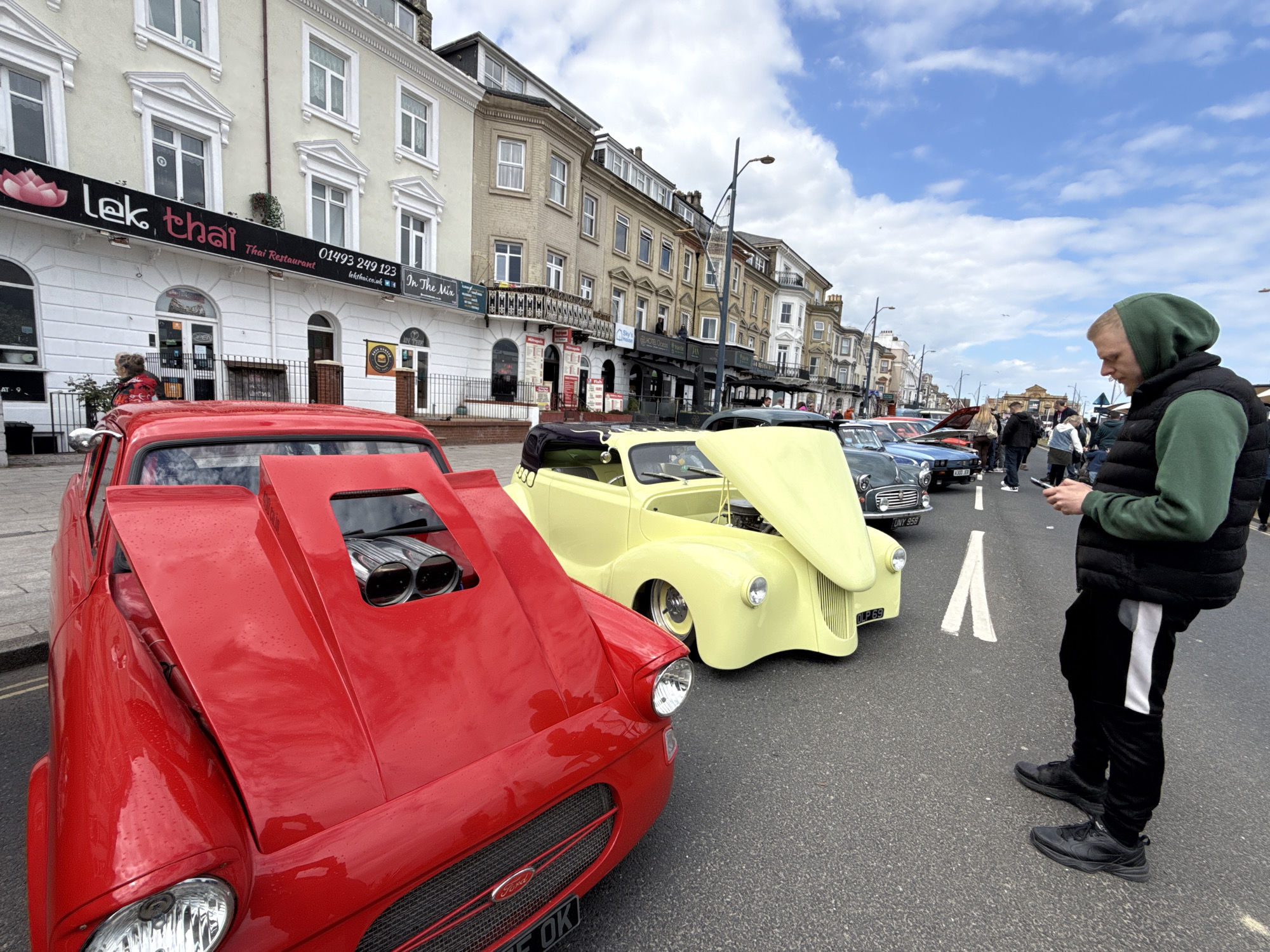 The image shows a street scene in Great Yarmouth, United Kingdom, featuring a row of vintage cars parked along a street, possibly for a car show or gathering. A red Ford car is in the foreground with its hood up, exposing the engine. Next to it is a light yellow car, also with its hood up. Several other vintage cars are lined up further down the street. A man stands to the right side of the image, looking at his phone. Behind the cars are various buildings with businesses; one is a Thai restaurant named "Lek, Thai". The street appears to be closed to normal traffic and has white directional markings on the pavement. The weather is partly cloudy. People can be seen walking in the background, indicating a public event.
