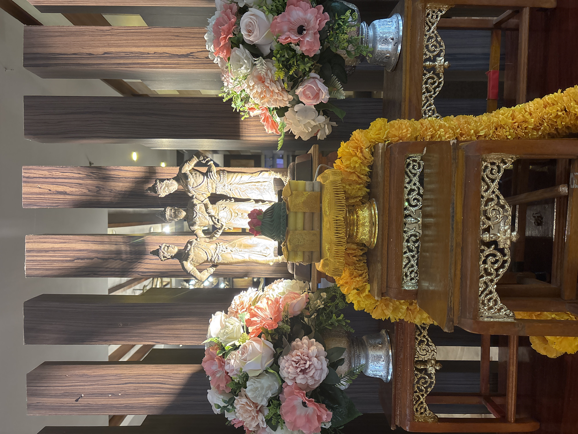 This image depicts an altar in Chiang Mai City Municipality, Thailand. Three bronze statues of figures in traditional attire stand against a backdrop of vertical wooden panels. In front of the statues, on a tiered wooden platform adorned with gold trim and a yellow floral garland, is a stack of objects topped with a green conical item. To the left and right of this central display are two silver vases overflowing with a floral arrangement of pink and white flowers. The scene is illuminated by soft lighting, casting shadows on the wooden panels and creating a serene atmosphere. The overall impression is one of cultural or religious significance, likely within a temple, shrine, or a dedicated space in a home or establishment.