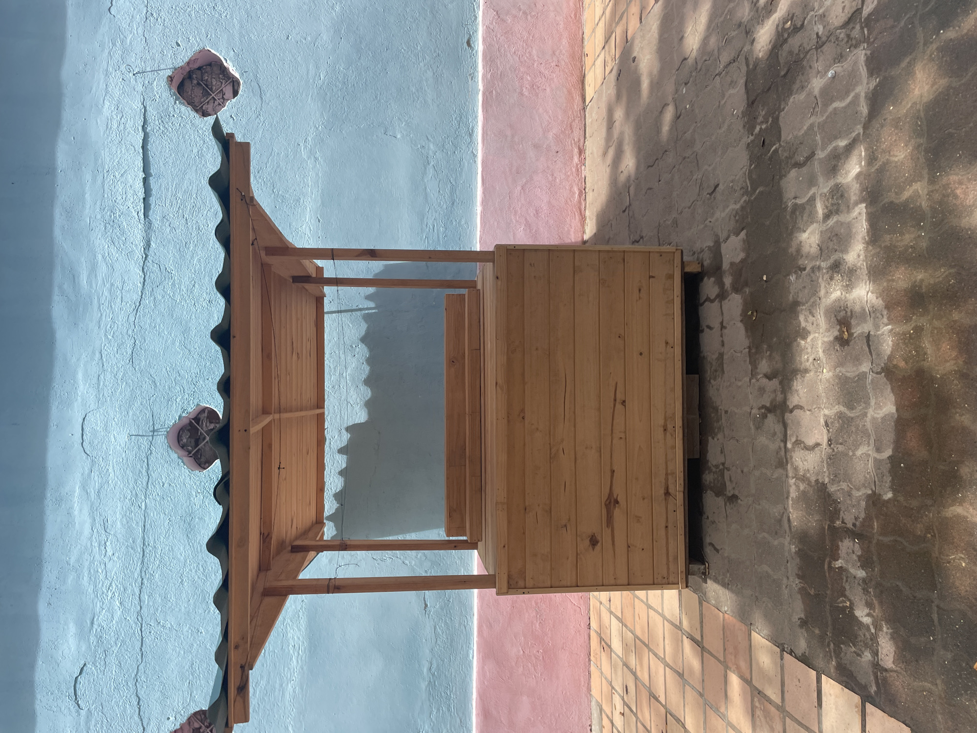 A wooden market stall with a dark, wavy roof stands in front of a blue and pink textured wall. The stall has a rectangular base constructed from horizontal wooden planks, topped with a wooden counter area. The roof is supported by four wooden posts. Two decorative, floral-shaped objects hang from the blue wall near the roofline. The ground in front of the stall is a mix of light brown square tiles and darker, patterned paving stones, some of which appear wet, casting shadows. The scene is outdoors, possibly in a city like El Valle del Espíritu Santo, Venezuela, given the architectural style and vibrant wall colors. There are no people or animals visible in the image.