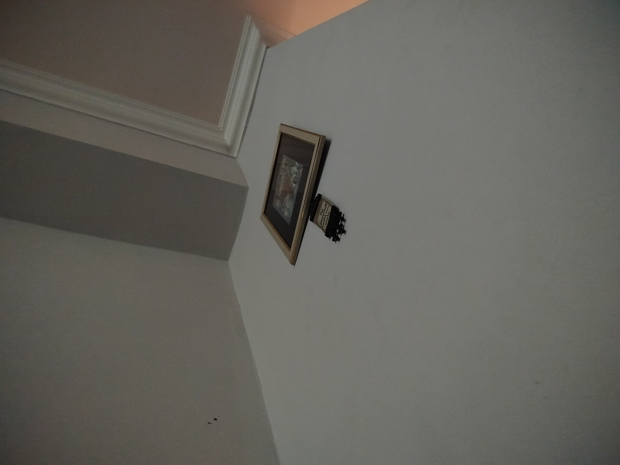 The image shows a framed picture hanging crookedly on a white wall. Attached to the picture frame is what appears to be a broken electrical outlet. The location is indoors. The city of Yerevan, Armenia is not discernible in the image.