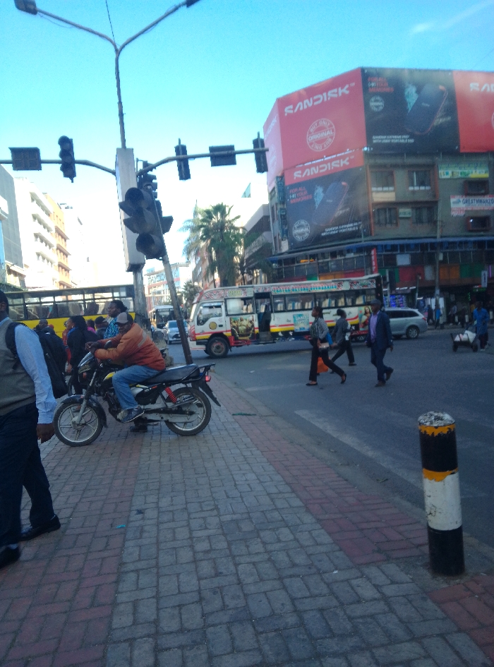 The image depicts a bustling urban street scene at an intersection in downtown Nairobi, Kenya, under a clear blue sky. A prominent multi-story building on the right features large red "SANDISK" billboards on its upper facade, advertising "GAMING EXTREME PRO" products and "ORIGINAL" branding.
In the foreground, a paved sidewalk with grey and reddish-brown brick patterns extends from the lower left towards the mid-right. A black motorcycle with red rear suspension accents is parked or stopped on this sidewalk, oriented towards the street. A male individual, wearing an orange jacket, blue jeans, and a light blue face mask, is seated on the motorcycle, facing left. To the immediate left, a person wearing a dark vest over a light blue shirt and dark trousers is partially visible, walking away from the camera on the sidewalk.
A busy asphalt road occupies the mid-ground. Traffic lights are visible overhead on a tall pole at the intersection. Vehicles include a colorful white "matatu" minibus with decorative artwork and text on its side (partially obscured, but "GREAT MWANZO" is discernible on a sign above a shop), and a silver sedan behind it, both moving or stopped in traffic. A large yellow bus is partially visible further left in the background.
Several pedestrians are crossing the road from right to left in the mid-ground: a woman in dark attire carrying a bag, followed by another individual, and a man in a dark suit. Additional pedestrians are visible on the far side of the street and near the bus/matatu. On the far right, a person is pushing a handcart. A black and white striped bollard with yellow at the top stands on the roadside in the lower right corner. Buildings of various heights and designs line the street.