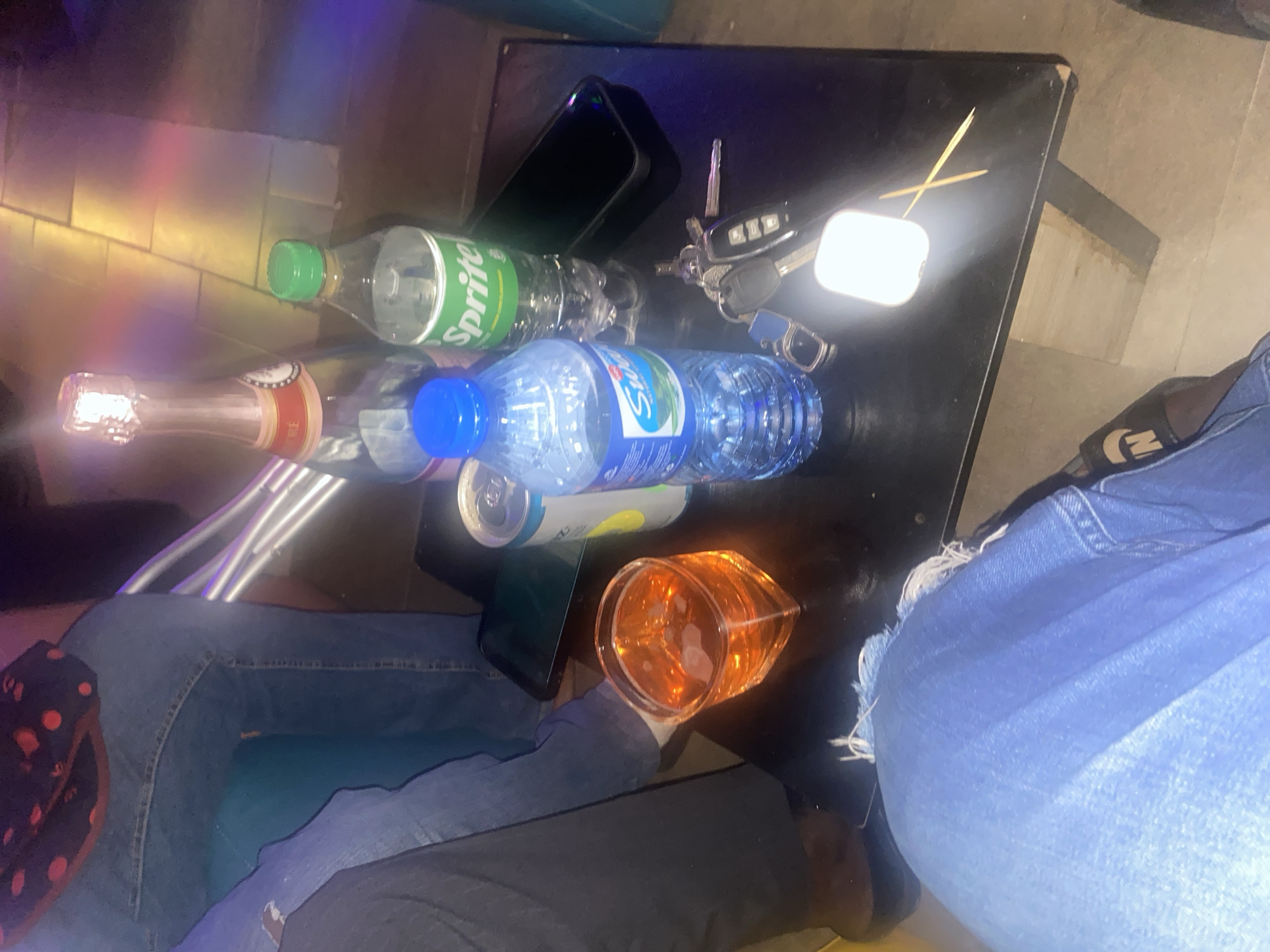 The image depicts a social gathering at a commercial establishment, likely a bar or lounge, in Jos, Nigeria, illuminated by dim ambient light and colorful projections. At least three individuals are partially visible, seated around a dark, rectangular table. In the immediate foreground, a person wears blue denim jeans with frayed fabric near the knee and a dark slide or sandal with an "N" logo on the right foot. To the upper left, another person's legs, also in blue jeans, are visible. On the left side of the table, a third person is partially seen, wearing dark trousers and a dark, patterned top.
The table's surface contains several objects: a champagne bottle with its cork wrapped in foil, a small plastic bottle of Sprite soda with a green cap, and a small plastic bottle of Swan Natural Spring Water with a blue cap. A square, clear glass holds an amber-colored liquid with visible ice cubes. A light-colored aluminum can is also present. Two dark-colored smartphones are placed face down on the table. A set of car keys, including a modern key fob, lies next to a small, rectangular white object emitting a bright light. Two wooden toothpicks are arranged in a cross pattern on the table. The background reveals light-colored flooring, possibly tiled, reflecting multicolored light, and vertical metallic structures.
