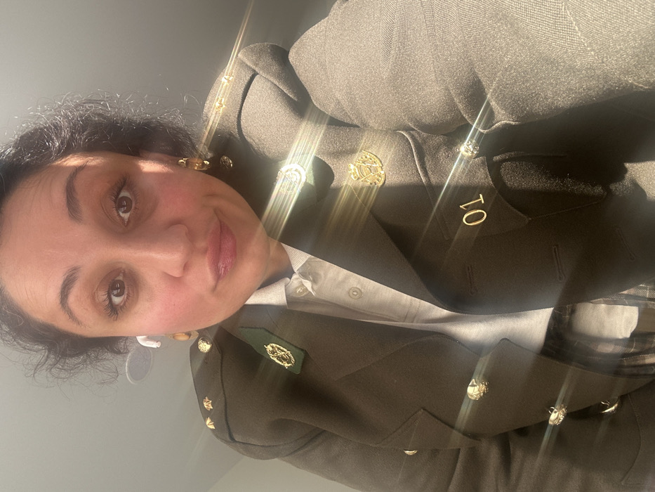 A woman with dark, curly hair and brown eyes looks directly at the camera, exhibiting a slight smirk. She is wearing a dark olive green, military-style double-breasted jacket over a light grey collared polo shirt. The jacket features three small gold stars on the left shoulder strap, a dark green collar patch with a gold emblem on the left, a gold circular crest on the right lapel, and the numeral "01" in gold on the right breast pocket flap. Gold-colored buttons are visible on the jacket's front closure. A white wireless earbud is present in her left ear, and she wears gold-colored dangling earrings with amber-like stones on both ears. The background is an indistinct, light-colored indoor setting. Multiple light flares are visible across the image. The image provides no visual cues to identify a specific location in Tbilisi, Georgia, or any other city.