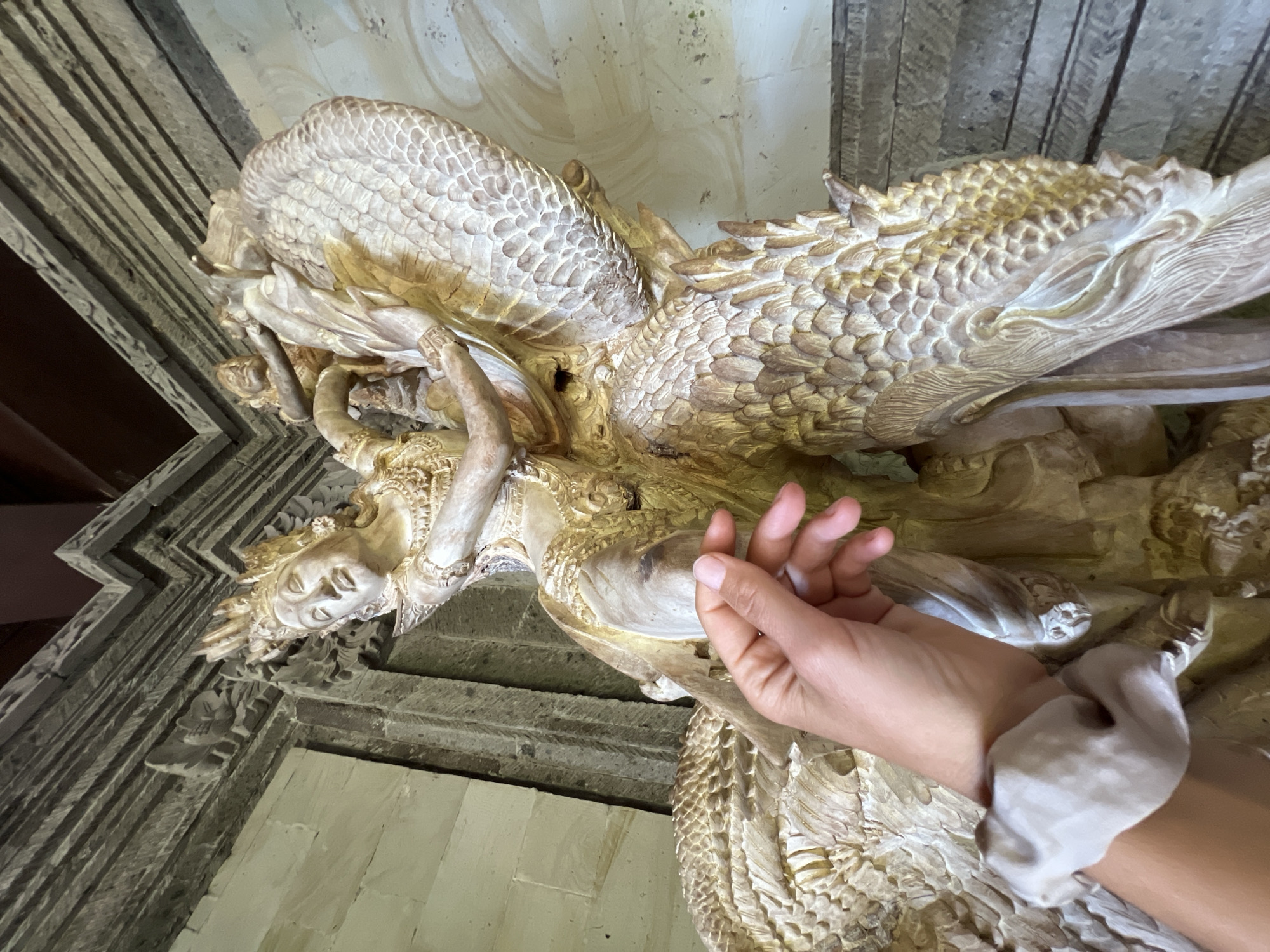 A low-angle, color photograph depicts a person's hand touching a carved stone sculpture on the exterior of a building in Kedewatan, Indonesia.
The sculpture is made of light-colored stone, appears old, and features an intricately detailed figure of a woman or angel, head down, and surrounded by large, scale-like wings that resemble those of a dragon or other mythical creature. The scales are finely carved, and the sculpture has a yellowed, aged appearance. The woman's face is serene, with her eyes closed.
A fair-skinned hand is visible in the foreground, with fingers gently touching the sculpture. A light gray scrunchie is around the wrist.
The building is constructed from stone blocks that are horizontally layered. There are carved stone decorations surrounding the edge of the structure.