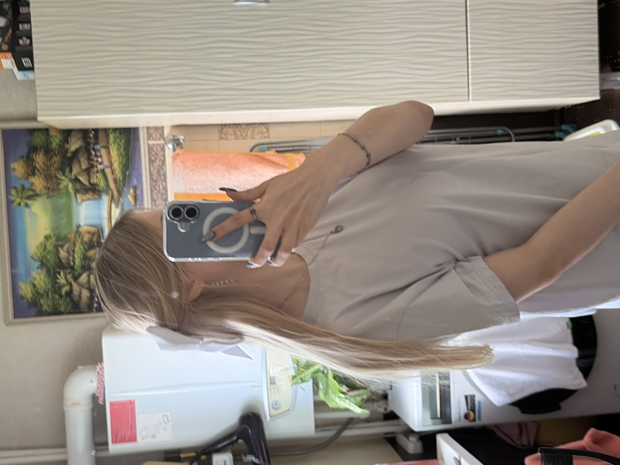 The image is a selfie taken in a bathroom. The camera is held by a person with long blonde hair, wearing a light grey t-shirt. The person's face is partially obscured by the phone. The bathroom contains a vanity with a cabinet above, a mirror reflecting a picture, and a white fixture of unknown purpose against the wall. The location is unknown.