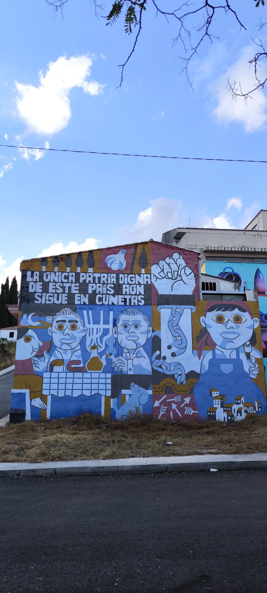 The image shows a mural painted on the exterior of a building in Fanzara, Spain. The mural is a colorful, stylized depiction of people and objects, with Spanish text across the top. The text reads "LA UNICA PATRIA DIGNA DE ESTE PAIS AUN SIGUE EN CUNETAS," which translates to "The only dignified homeland of this country still remains in ditches."
The mural features several distinct panels. On the left, two men with exaggerated features are seated at a table covered with a checkered tablecloth. One man is eating, and the other appears to be holding something, possibly a cigarette. A small dog sits at their feet. To the right of these figures, a large, stylized fist is depicted, signifying a struggle or protest. Further to the right, a woman in overalls and a bandana is shown holding a tool, with a collection of small houses below her. Between the figures, there are abstract elements including chains and arrows.
The building itself is made of brick and painted with various colors, including red, yellow, blue, and white. The sky above is blue with scattered white clouds. Trees are visible on the left side of the image, and a portion of another building can be seen in the background. The mural extends along the length of the building's facade. The foreground shows a patch of dry grass and a paved road with a sidewalk. The signature "ELIAS TAMO" is visible on the lower right section of the mural.