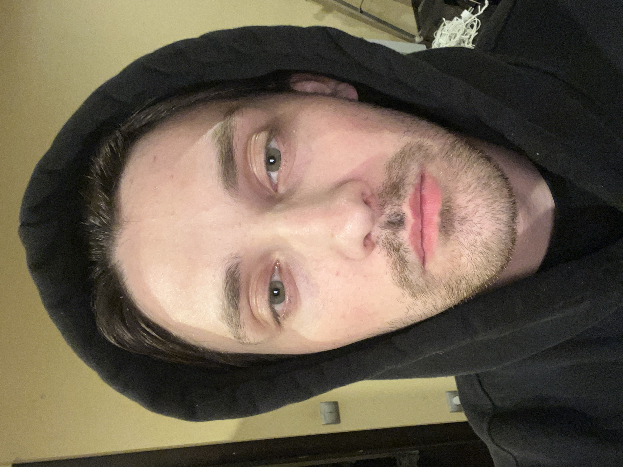 The image features a young man with light skin, dark hair, and light-colored eyes, appearing to be green or hazel. He is wearing a black hooded sweatshirt (hoodie), with the hood pulled up, framing his face. The image is rotated 90 degrees to the left, showing his head tilted to the right.
The man has short, dark stubble on his chin, jawline, and upper lip. His lips are slightly parted, and he has a neutral or contemplative expression as he looks directly into the camera. There are subtle highlights on his forehead and the bridge of his nose, suggesting indoor lighting. His dark hair is visible at the edges of the hood.
The background is simple and out of focus. To his upper left (due to the rotation), a plain light beige or tan wall is visible. To his lower right, a darker, indistinct area contains what appears to be tangled white material, possibly wires or string. A light switch or outlet is visible on the wall behind his lower right eye. No text is visible in the image. The scene suggests an indoor setting, likely taken in Ореховка, Russia.